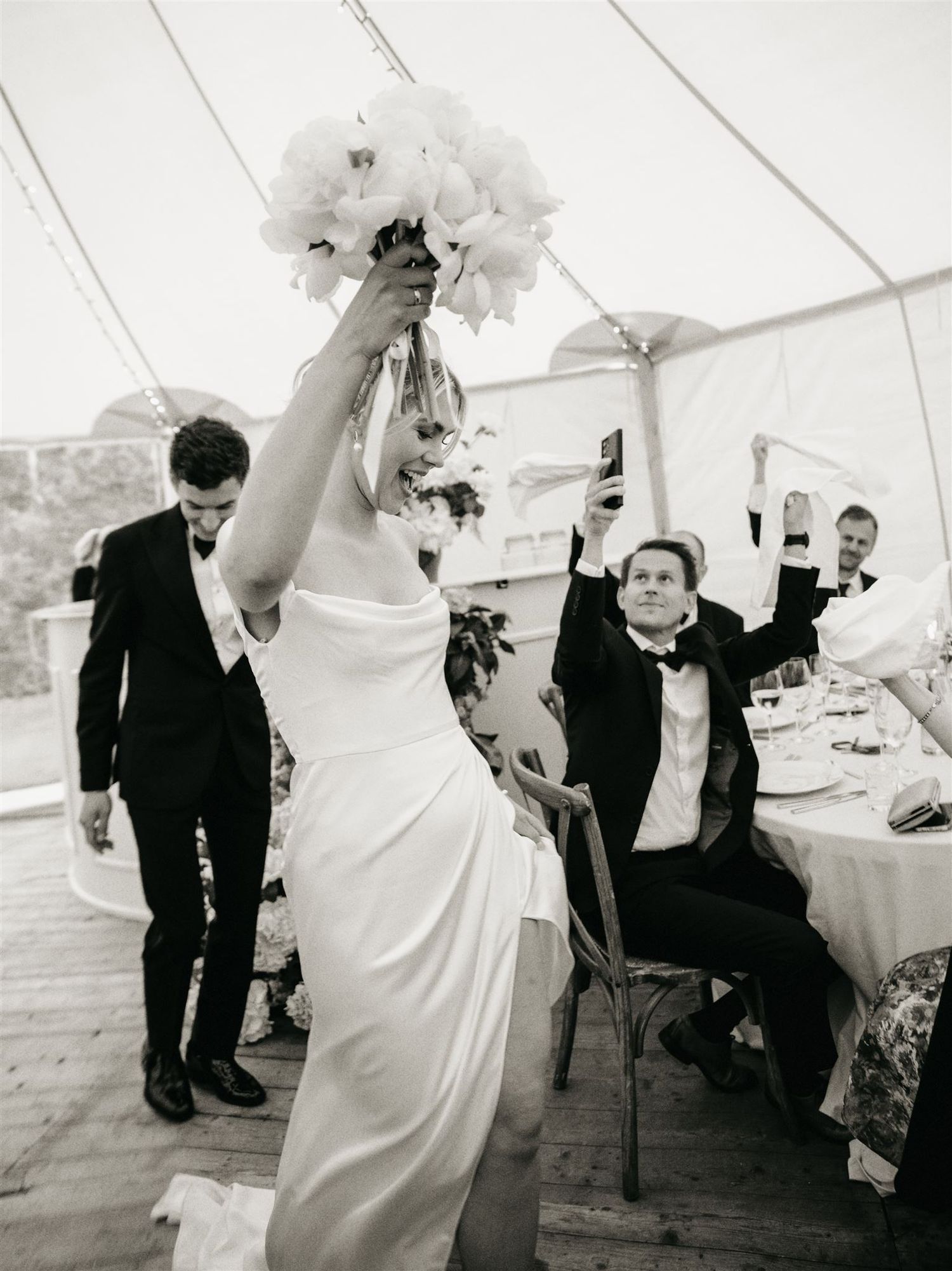 Bride raises bouquet of white peonies triumphantly at wedding reception in black and white.