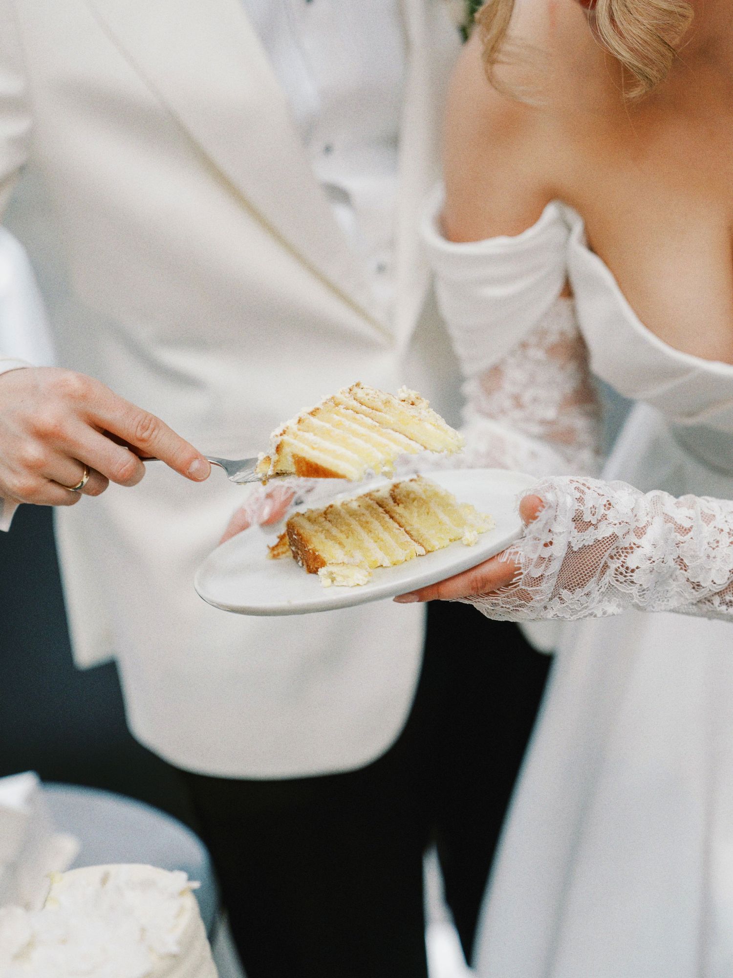 Bride and groom cutting a white layered wedding cake, with the bride in a lace gown holding a plate.