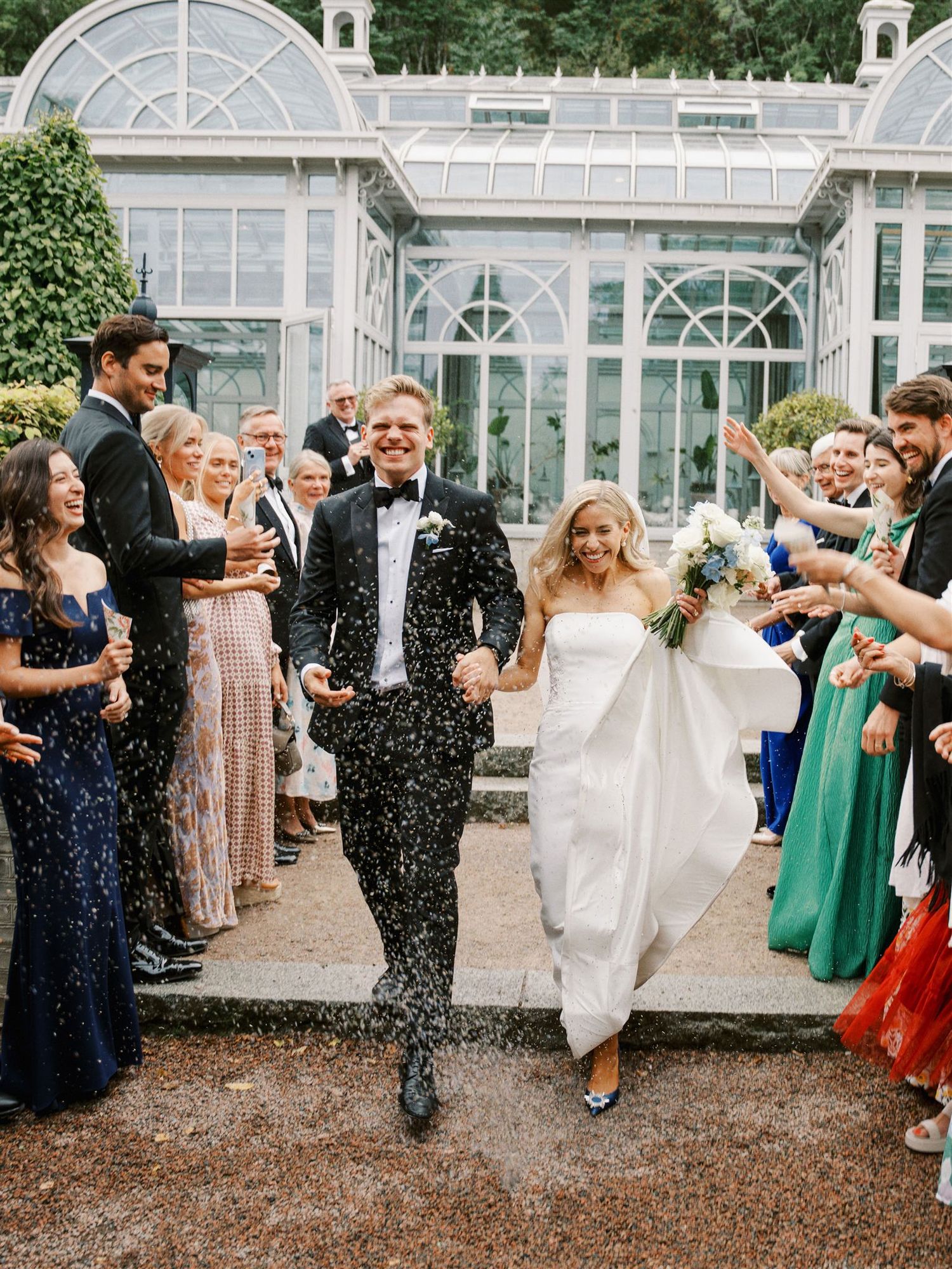 Joyful bride and groom exit greenhouse venue through cheering guests tossing confetti celebration.