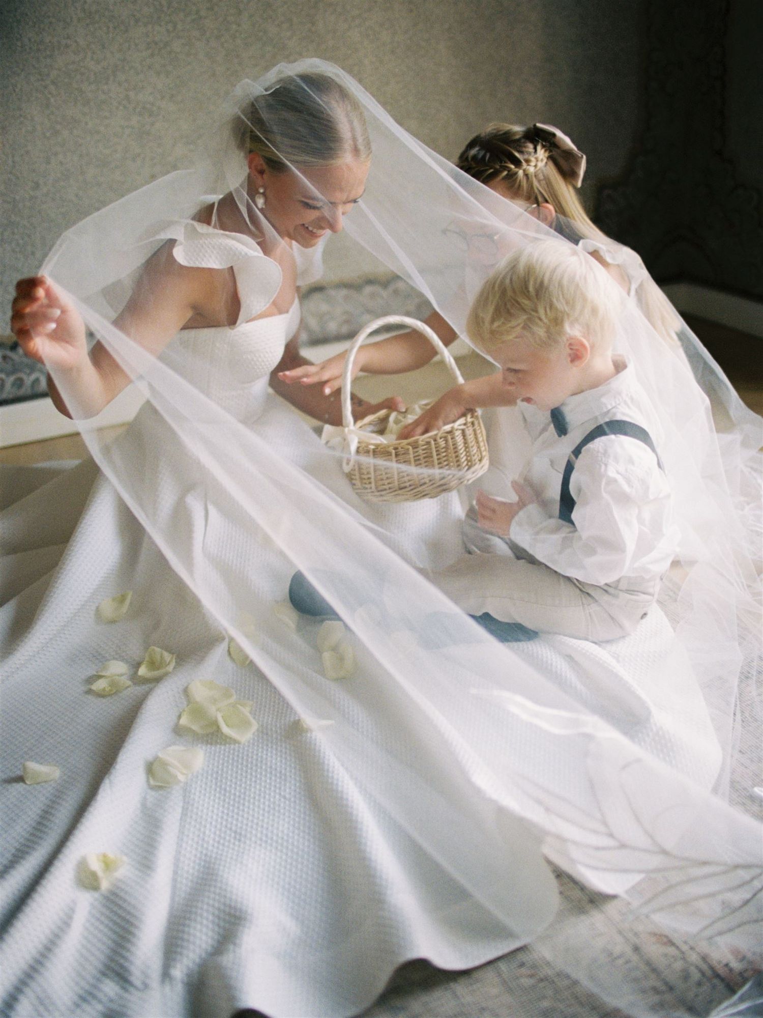 Bride in white gown and veil shares a tender moment with young flower girl and ring bearer on wedding day.