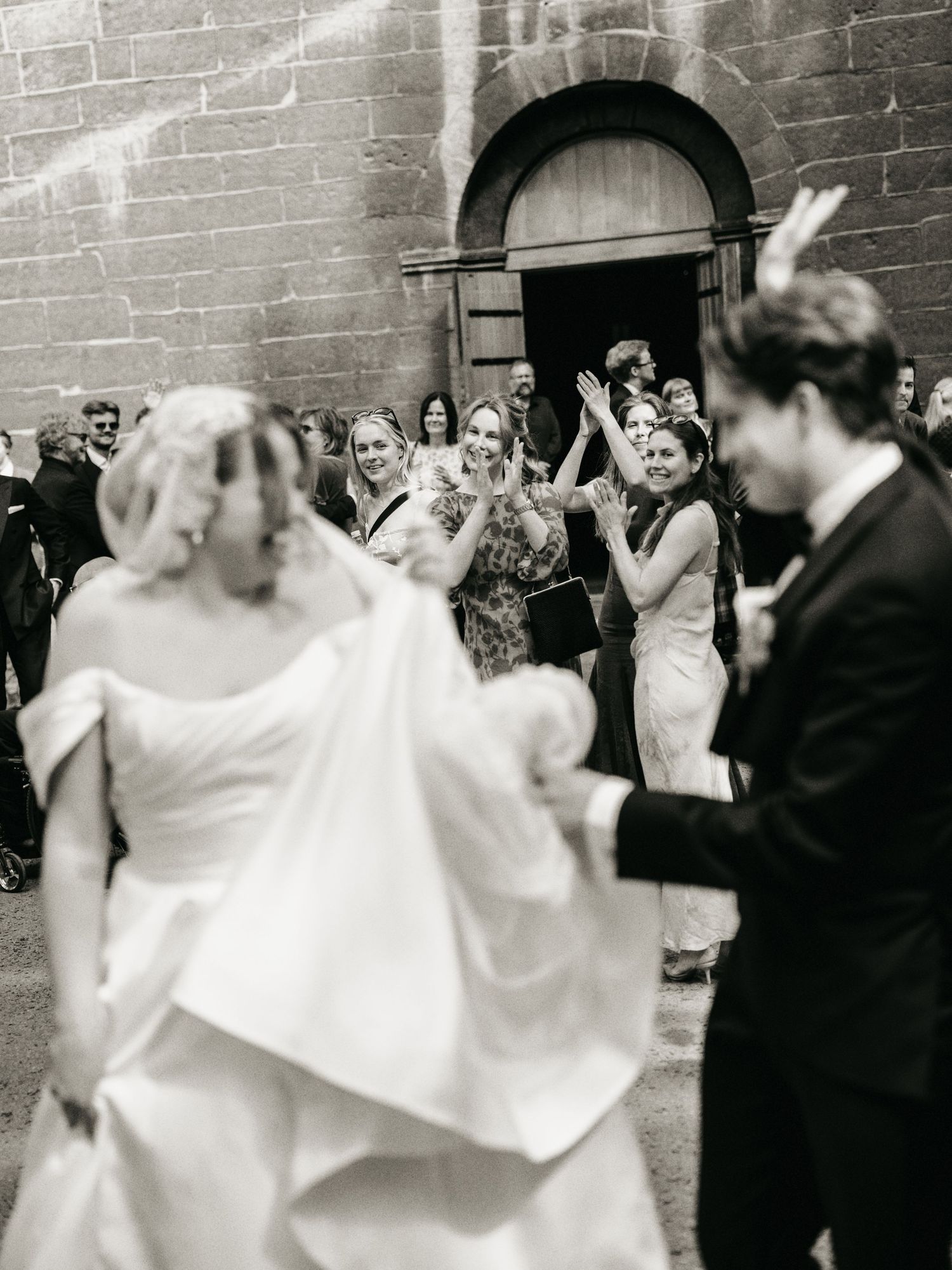 Black and white photo of a bride and groom exiting a church as guests clap and celebrate their wedding ceremony.