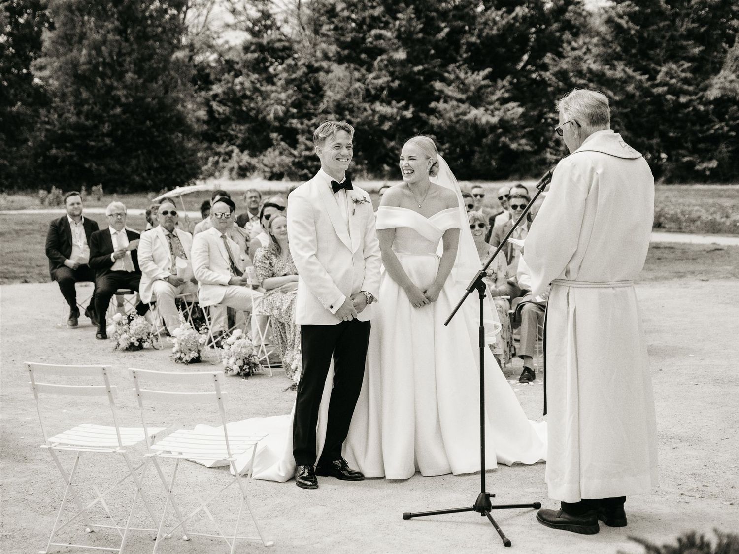 Black and white photo of bride and groom exchanging vows with officiant at outdoor wedding ceremony.