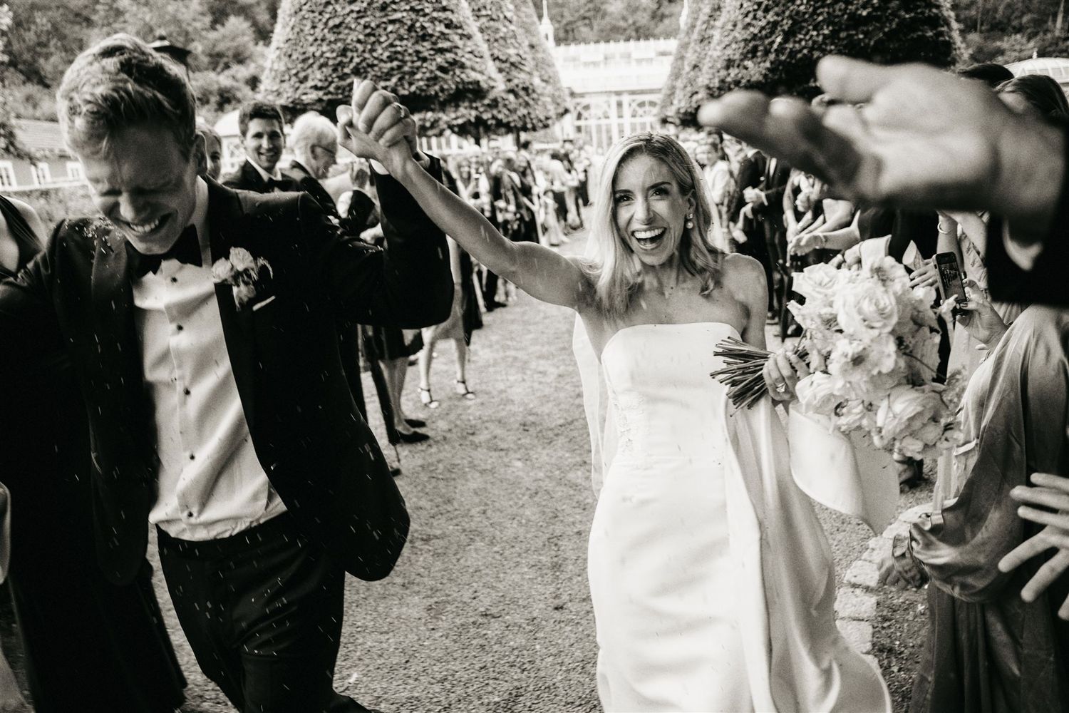 Black and white photo of joyful bride and groom celebrating with guests outdoors after wedding ceremony.