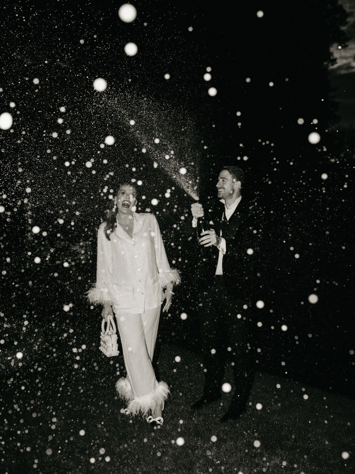 Bride and groom celebrate at night with champagne spray creating magical bokeh effect in black and white.