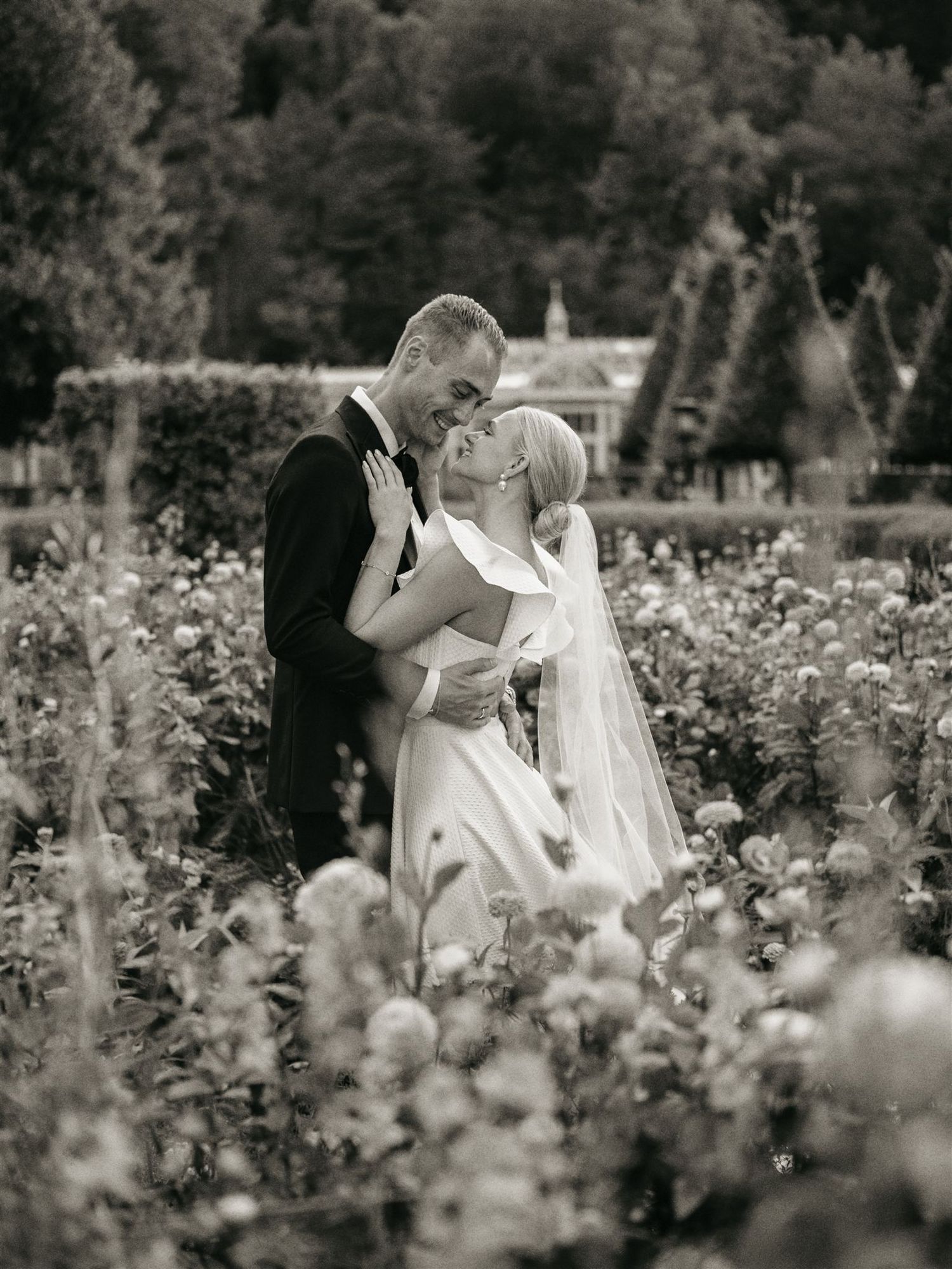 Bride and groom embrace romantically in a lush garden surrounded by blooming flowers in black and white.