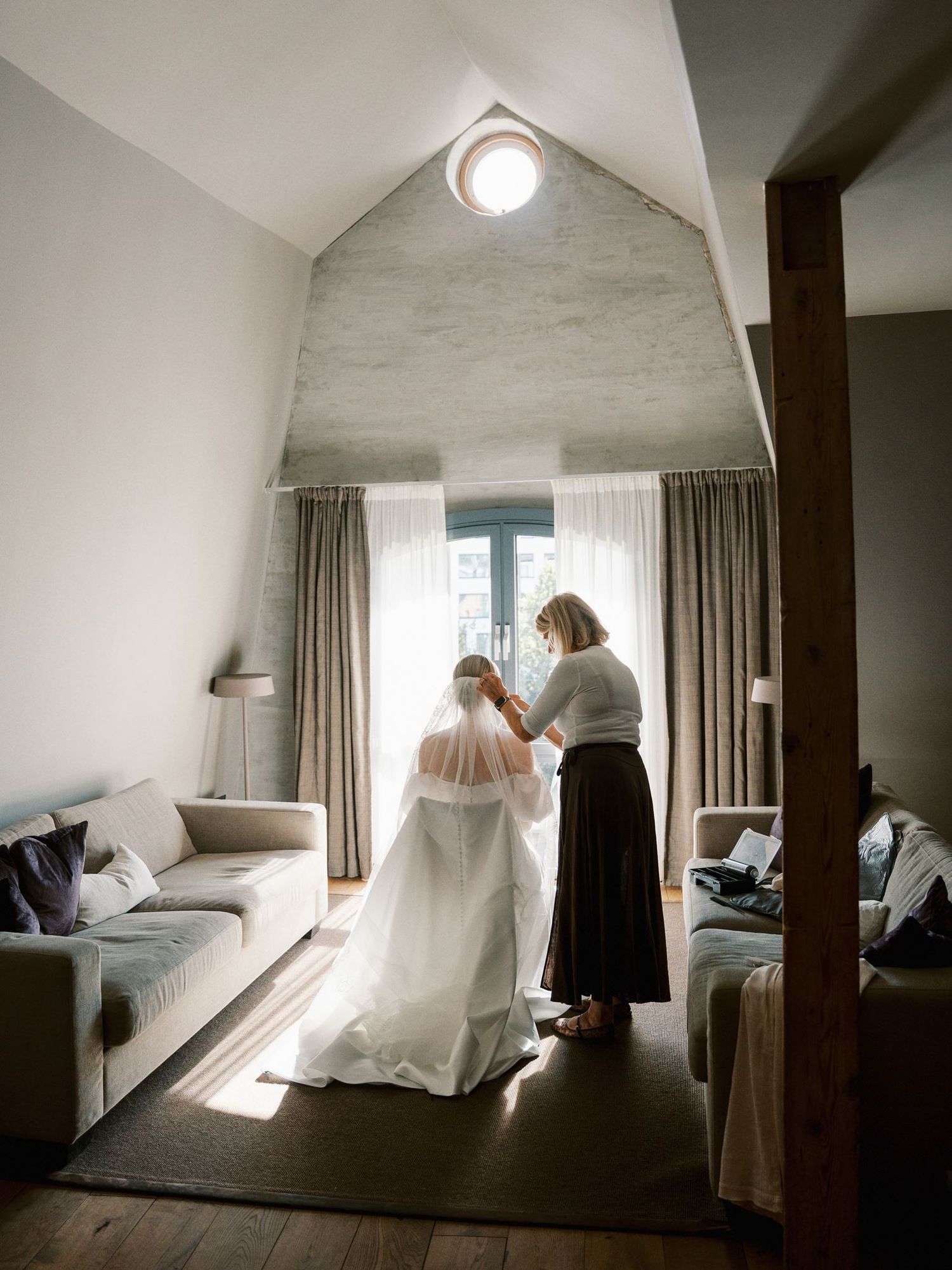 A bride getting her veil adjusted by a helper in a modern, light-filled bridal suite before the wedding ceremony.
