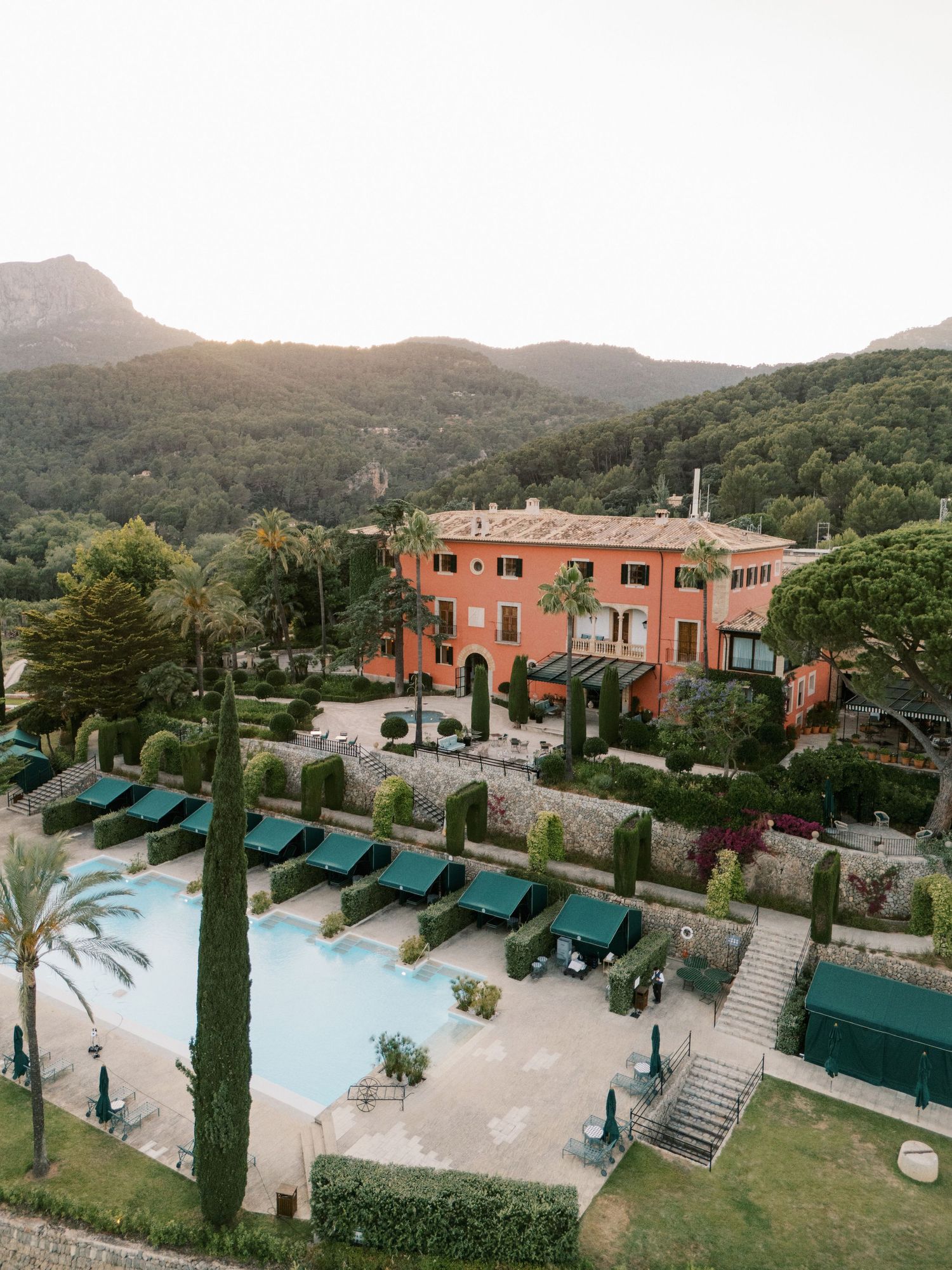Aerial view of a terracotta Mediterranean villa with pool, sun loungers, and lush mountain backdrop.