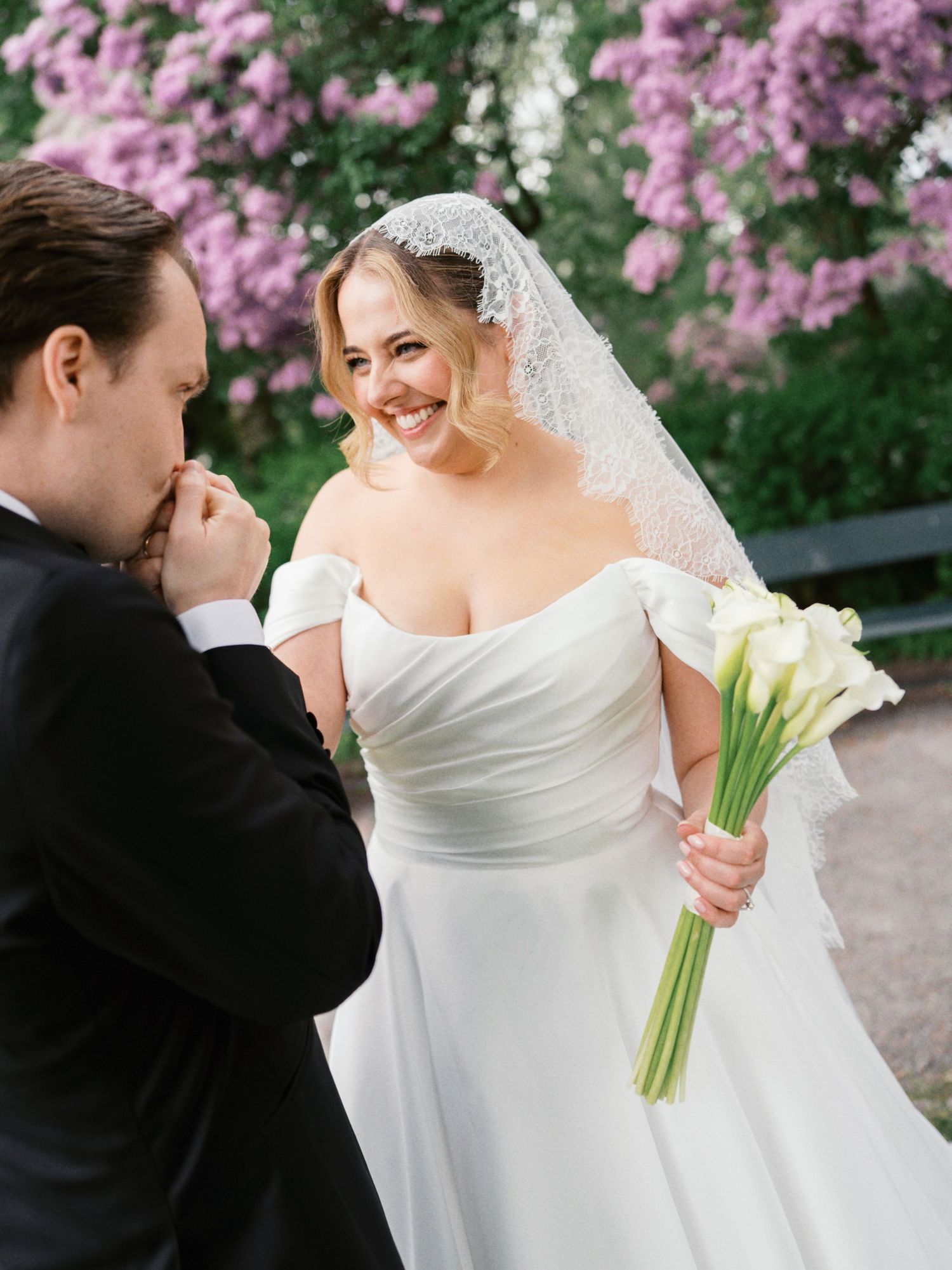 Bride in off-shoulder white gown holds calla lily bouquet as groom in tuxedo kisses her hand near blooming purple flowers.