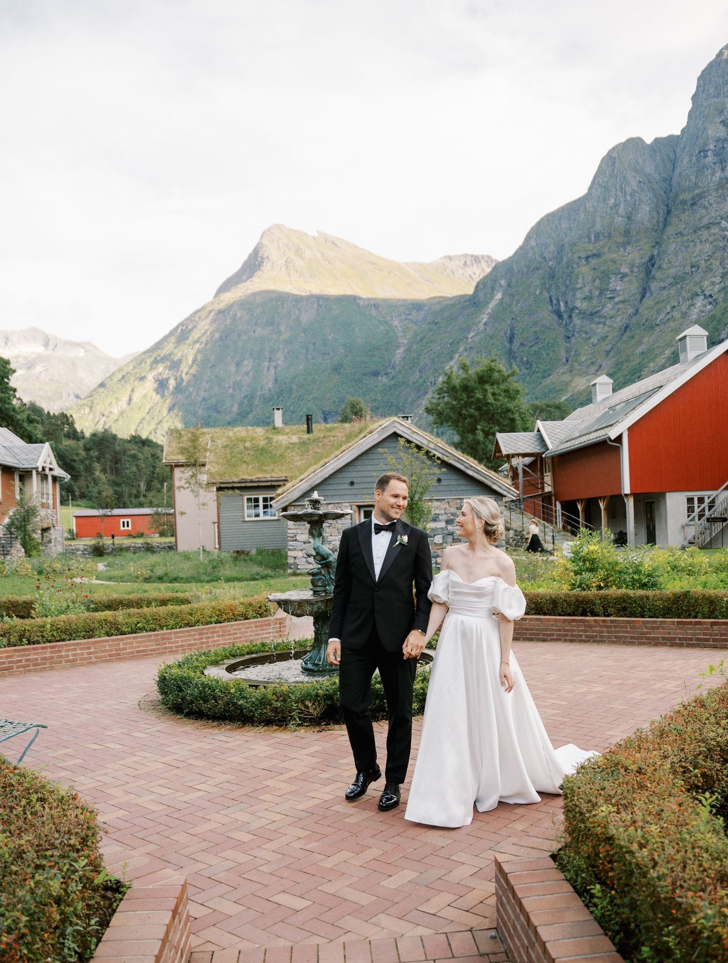 Bride and groom walking in a garden courtyard with grass-roofed buildings and dramatic Norwegian mountains in the background.
