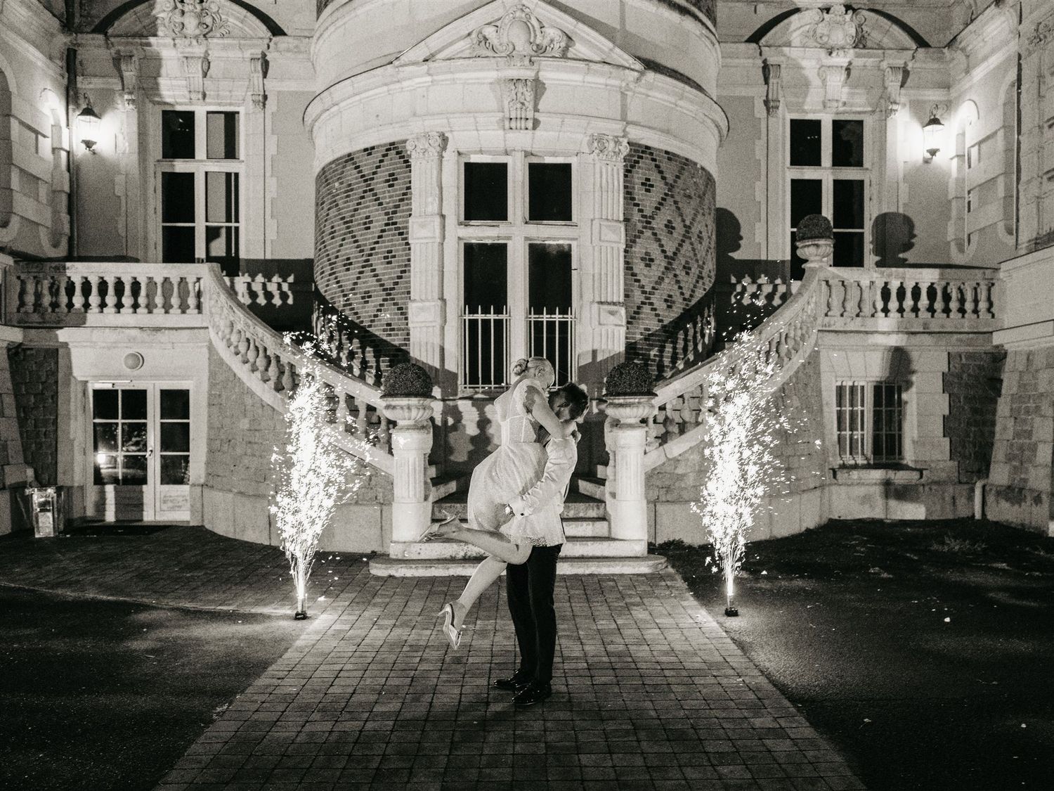 Bride and groom embrace dramatically with sparklers at night outside illuminated chateau in black and white.