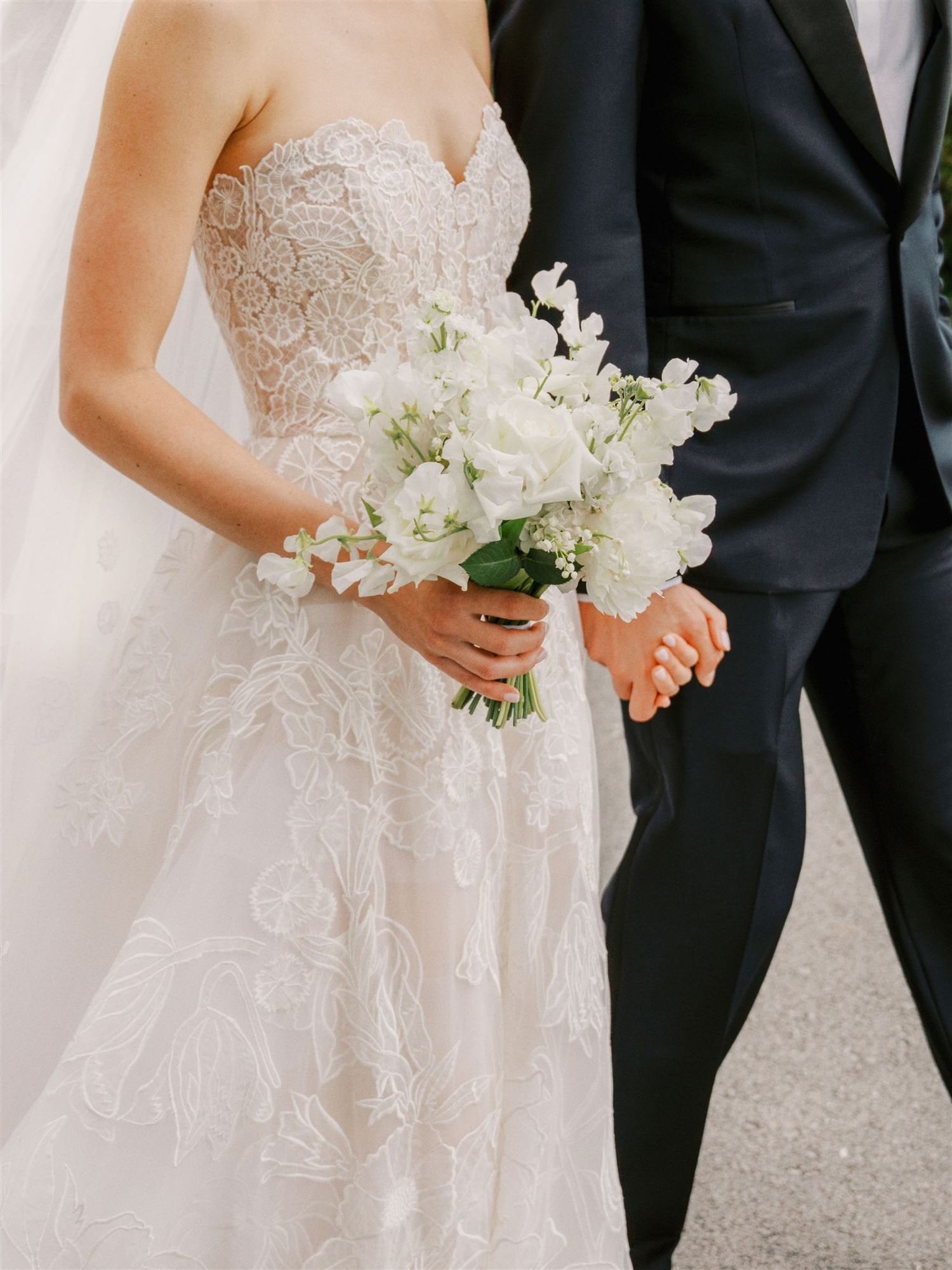 Bride in strapless lace wedding gown holding elegant white floral bouquet beside groom in dark suit.