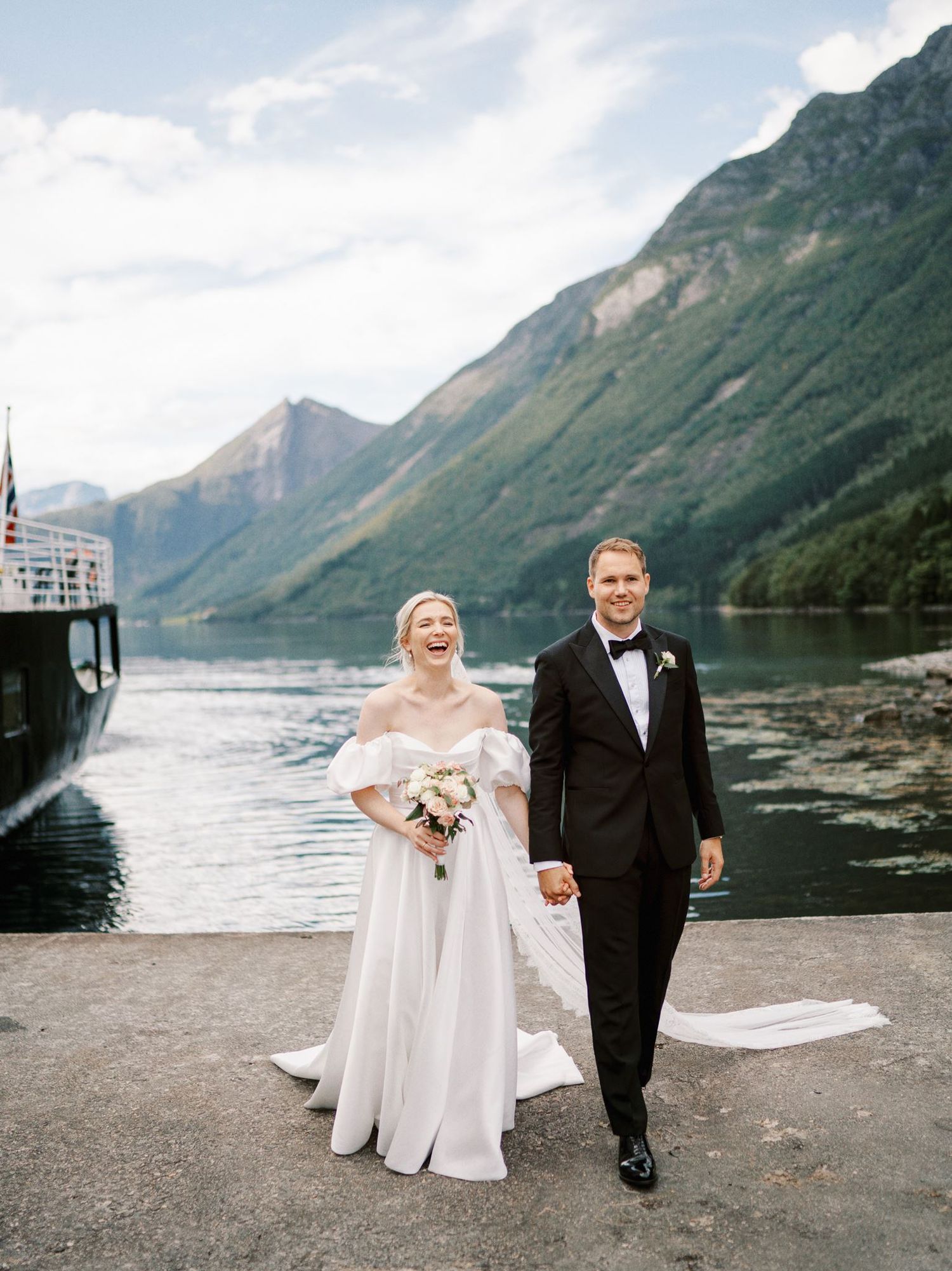 A joyful bride and groom walk hand-in-hand on a dock with stunning Norwegian fjord mountains in the background.