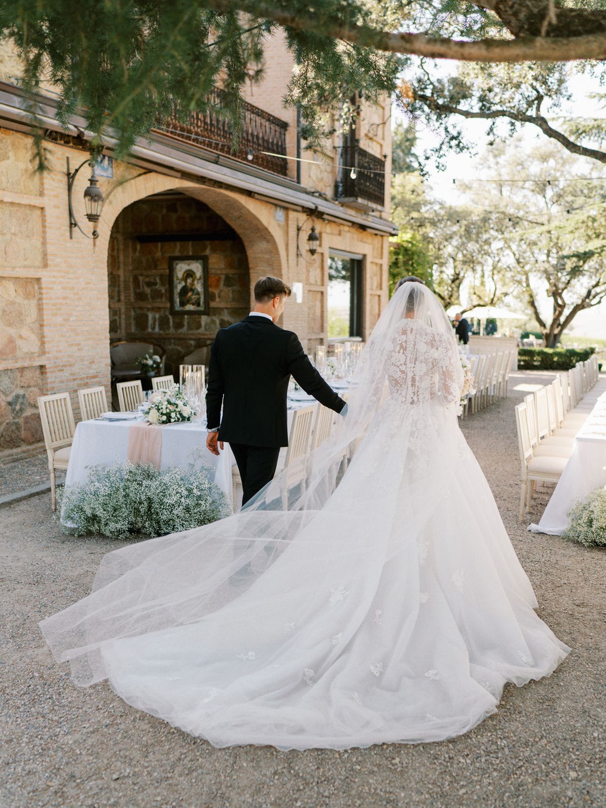 Bride in lace gown with cathedral veil and groom walk toward elegant outdoor wedding reception venue.