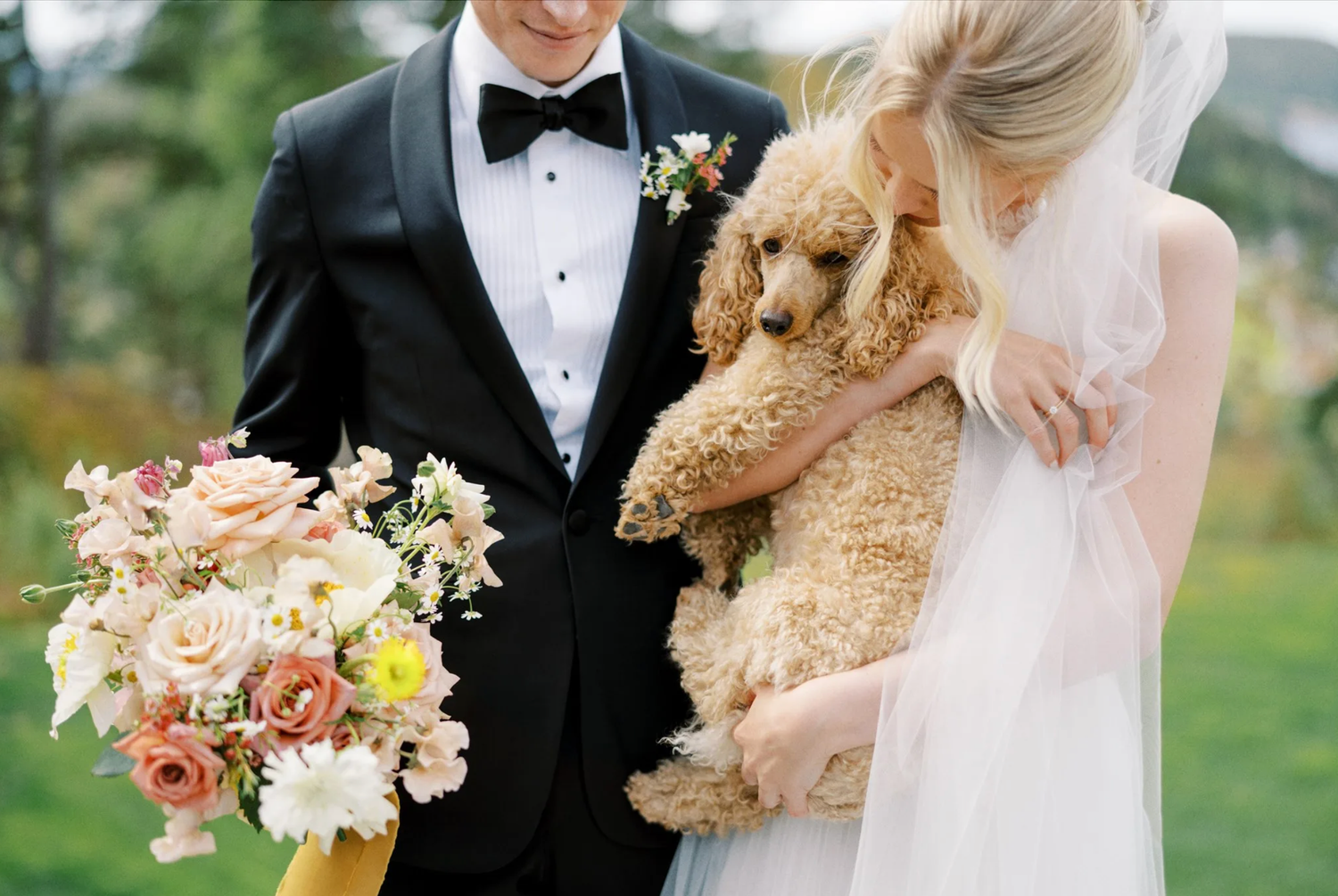 Groom in tuxedo and bride in white gown holding adorable fluffy apricot poodle and floral bouquet.