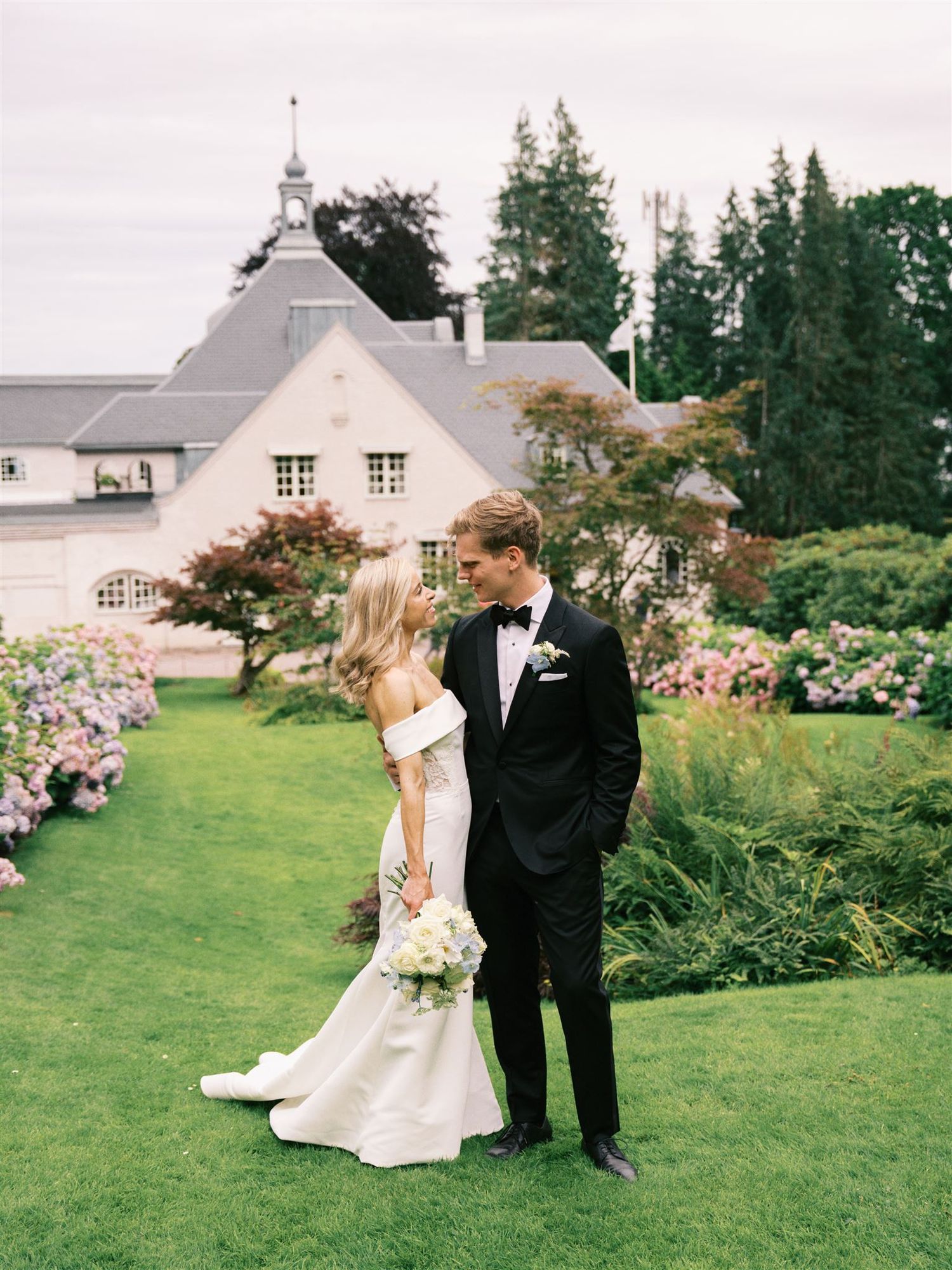Bride and groom sharing romantic moment in lush garden with hydrangeas beside elegant manor house.