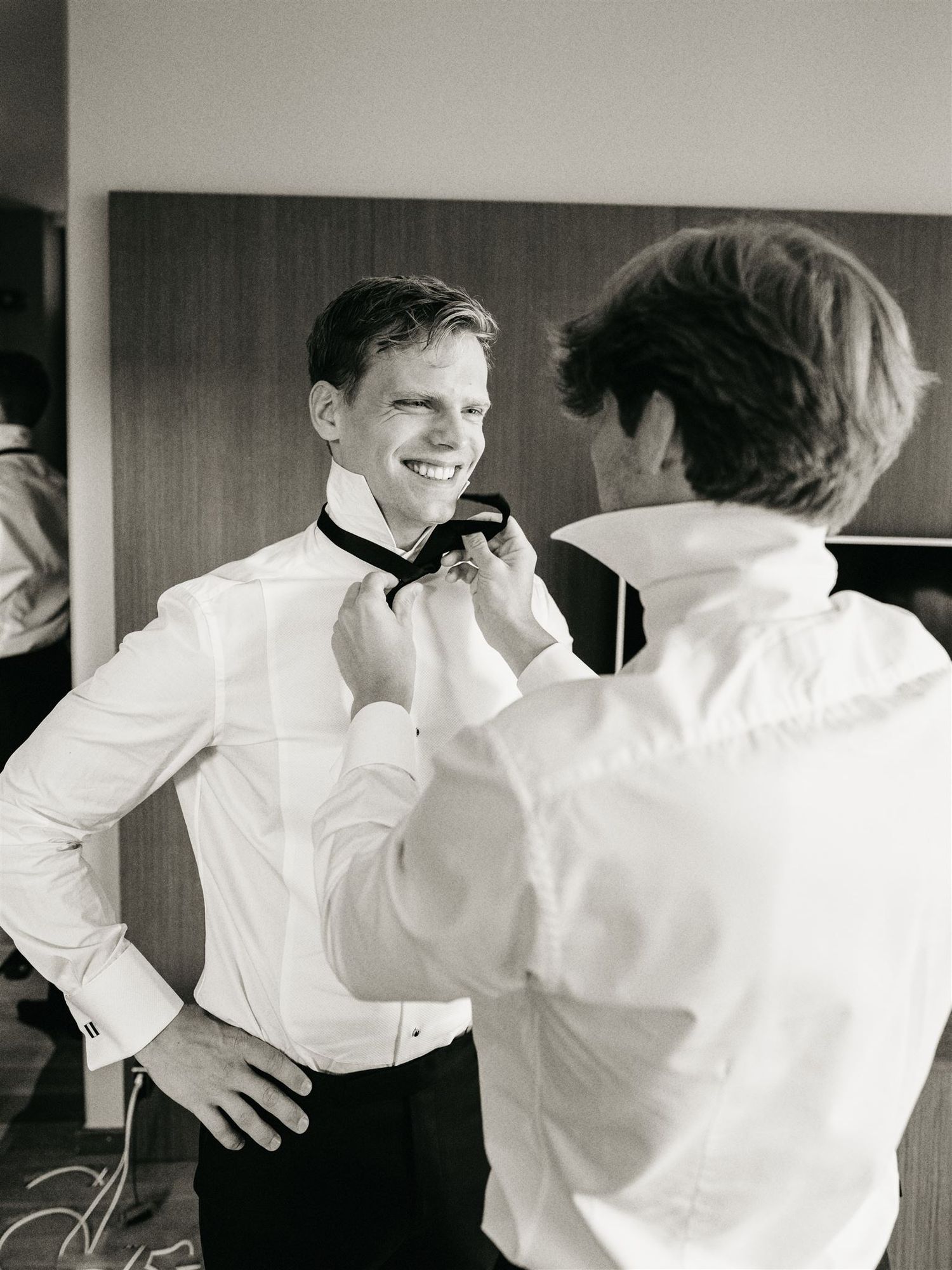 Smiling groom having his black bow tie adjusted by a groomsman in black and white photo.