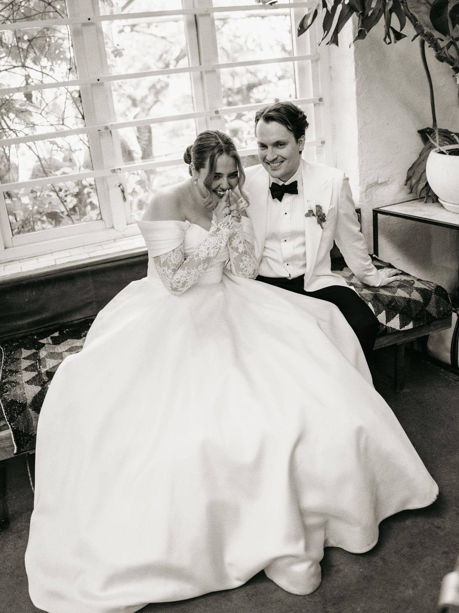 Black and white photo of a bride in an off-shoulder gown and groom in white tuxedo sitting together, laughing joyfully.