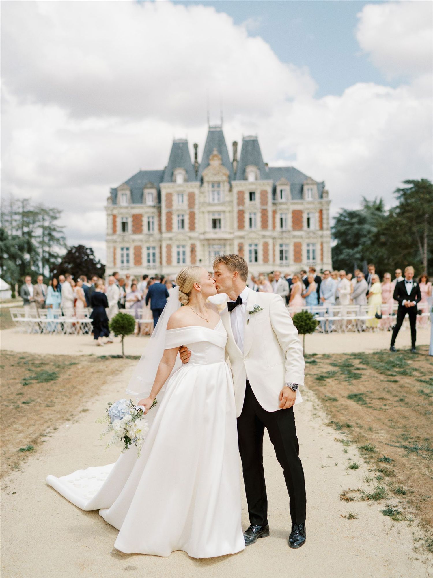 Bride and groom sharing a kiss in front of elegant French chateau with wedding guests behind them.