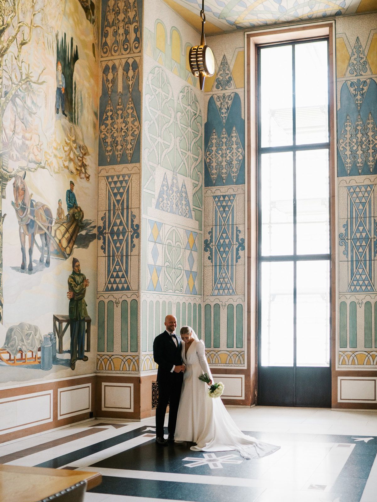 Bride and groom posing in stunning ornate hall with colorful murals and tall windows.