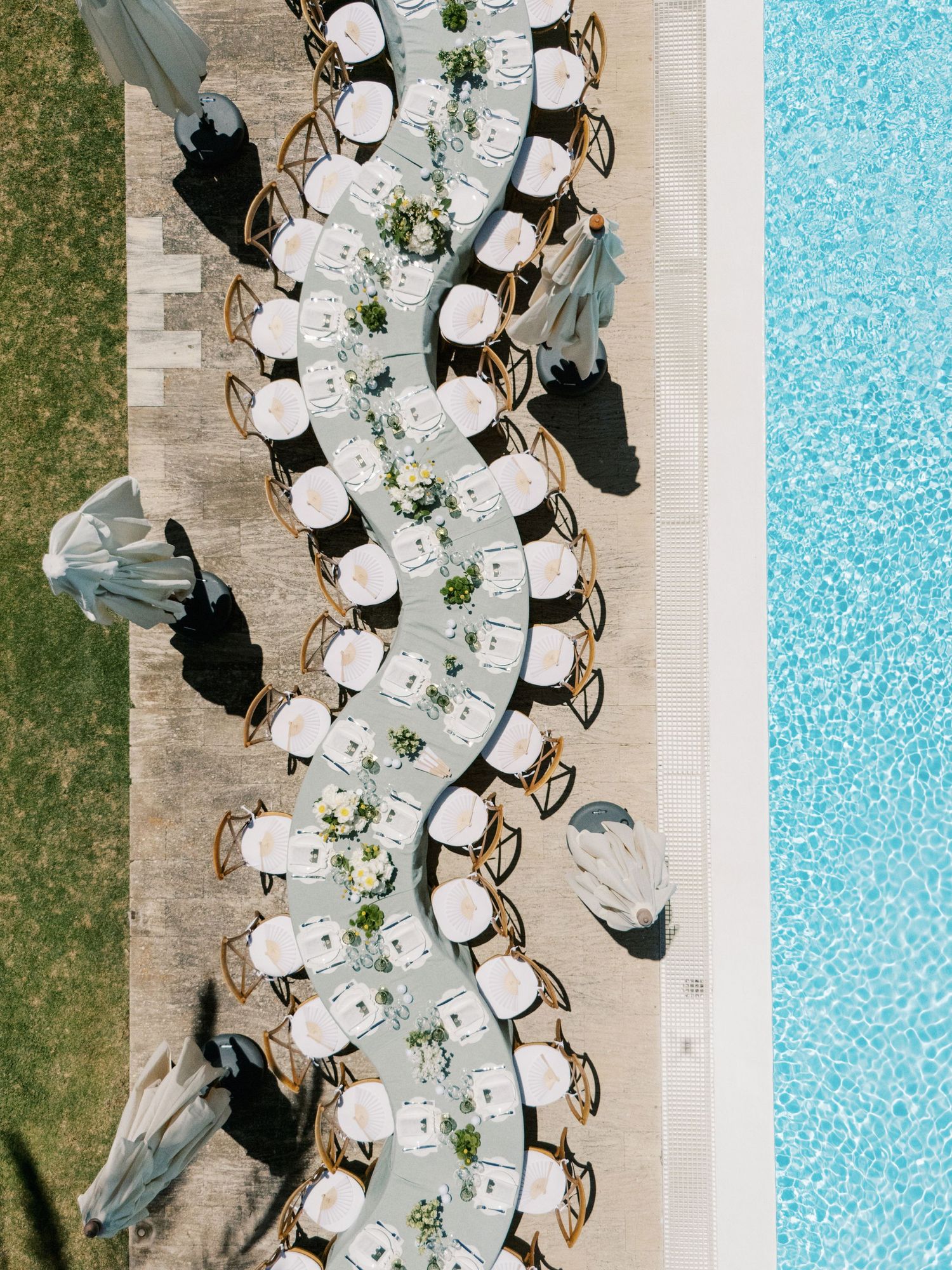 Aerial view of an elegant outdoor wedding reception table set poolside with white linens and floral centerpieces.