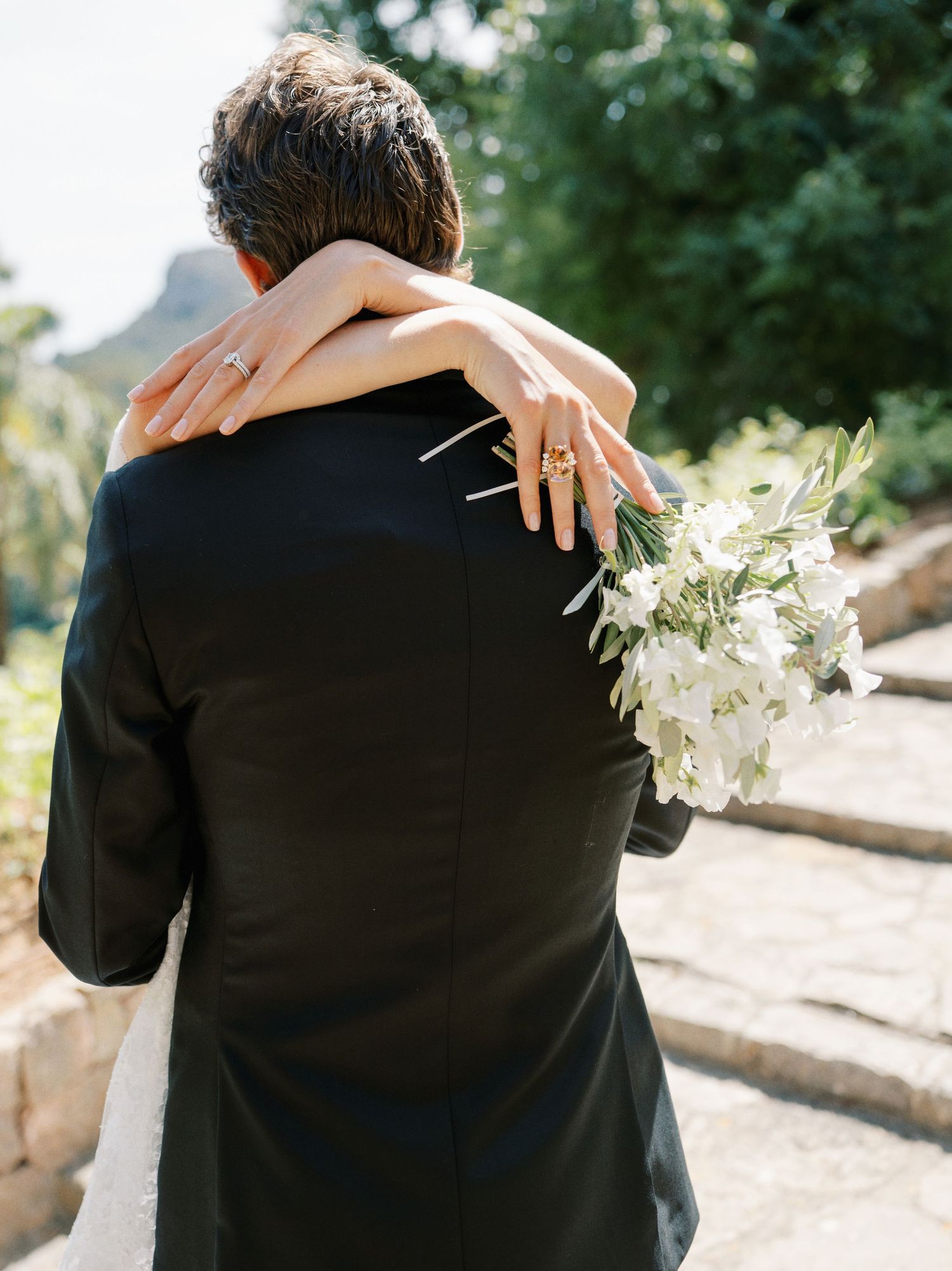 Bride holding white floral bouquet embraces groom in black suit from behind at outdoor wedding venue.