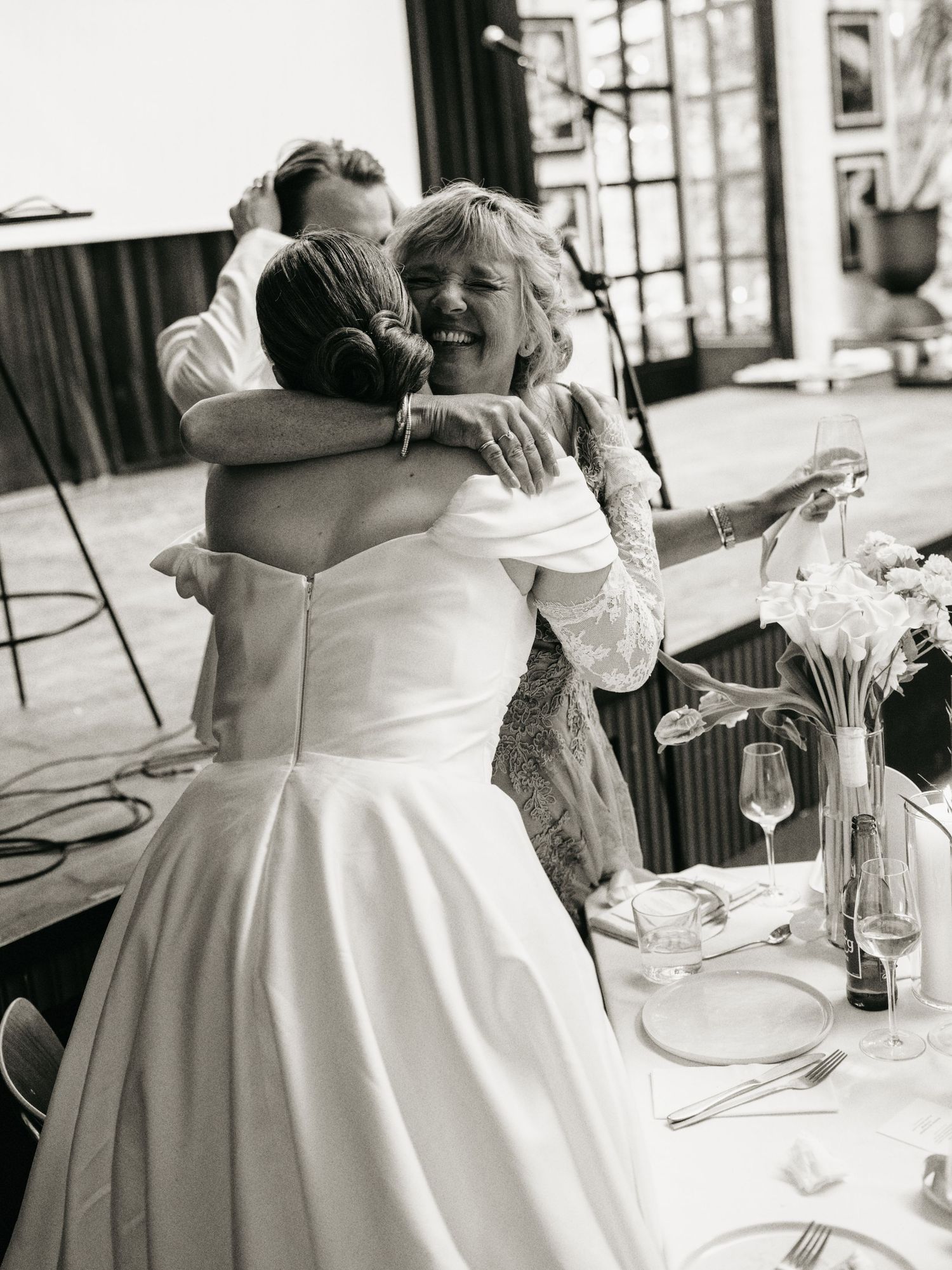 Black and white photo of a bride in a white gown hugging a smiling woman at a wedding reception.