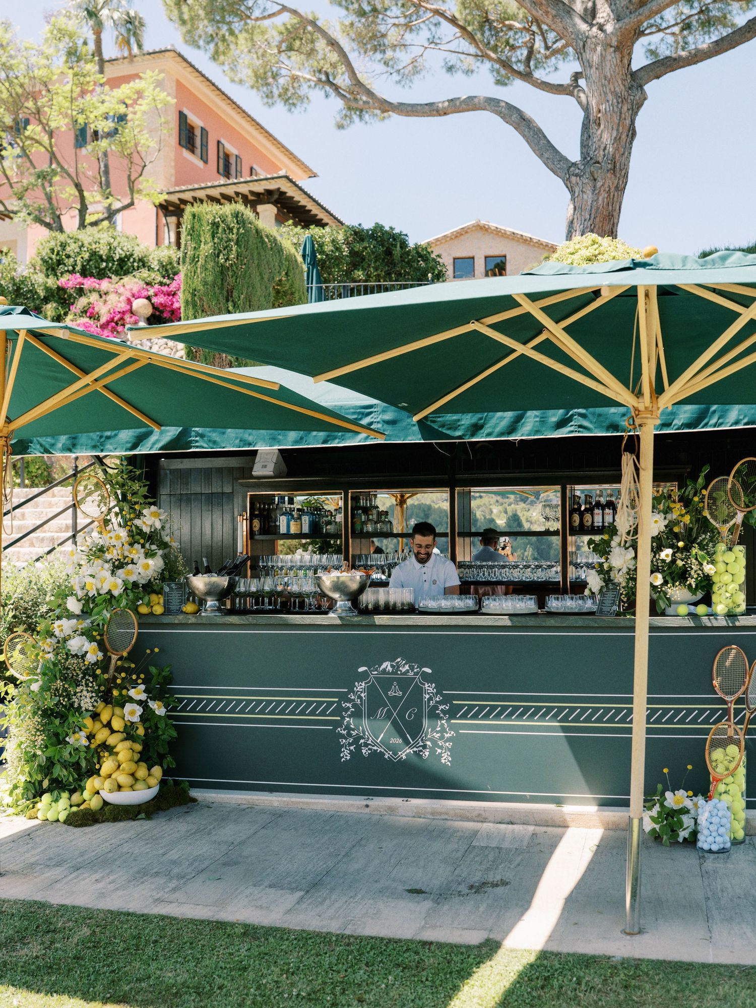Elegant outdoor bar with green and gold umbrellas, floral decor, and a bartender serving drinks at a garden event.