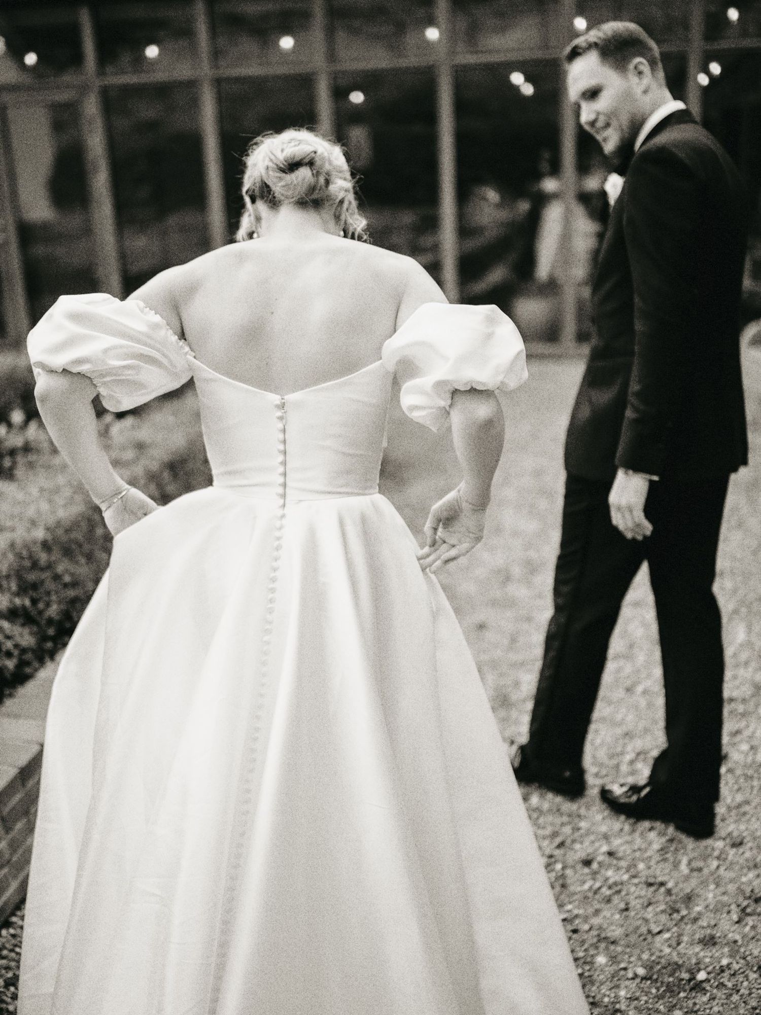Black and white photo of bride in off-shoulder gown with puff sleeves and button back, standing with groom in tuxedo.
