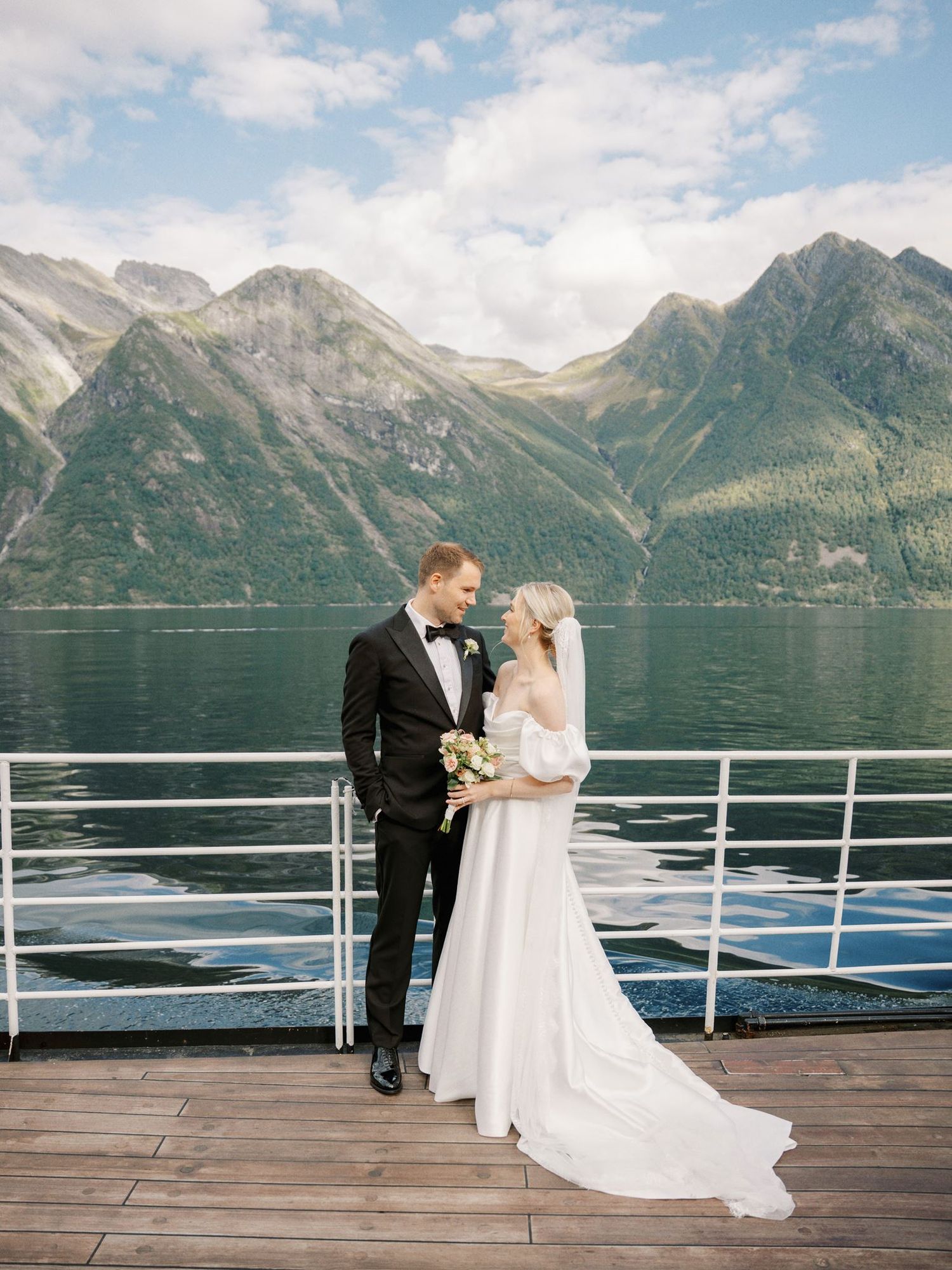 Bride and groom share a romantic moment on a wooden deck overlooking a stunning Norwegian fjord with mountain backdrop.