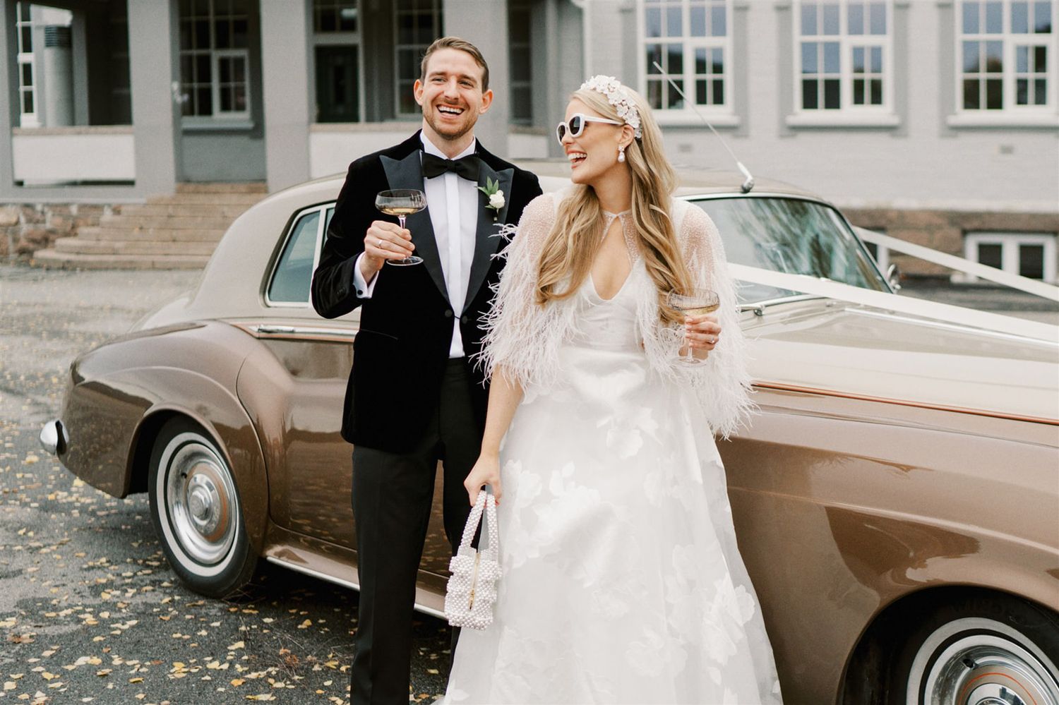 Bride and groom toasting with champagne beside a vintage tan car on their wedding day.