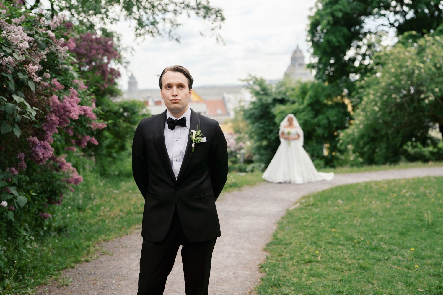 Groom in black tuxedo stands on garden path with bride in white gown blurred in background at outdoor wedding.