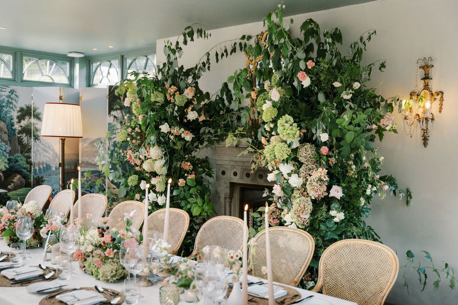 Elegant wedding reception with lush floral arch over fireplace, white roses, greenery, candlelit table, and French chairs.
