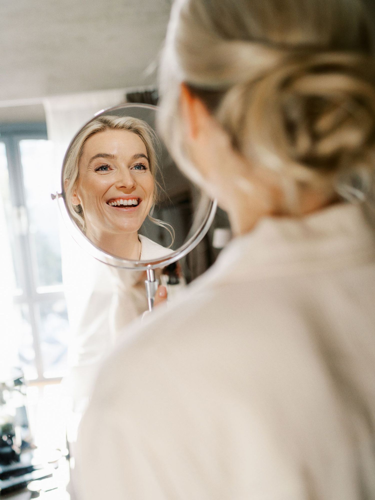Smiling bride admires her wedding makeup and updo hairstyle in a round mirror while getting ready.