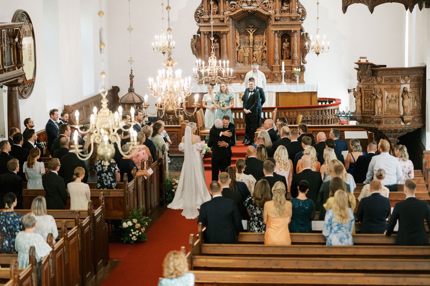 Wedding ceremony in an ornate church with guests seated in wooden pews, bride and groom at the altar under chandeliers.
