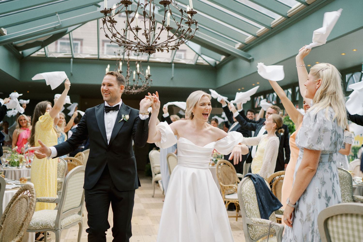 Bride and groom walk joyfully through guests waving paper at their elegant indoor wedding reception.