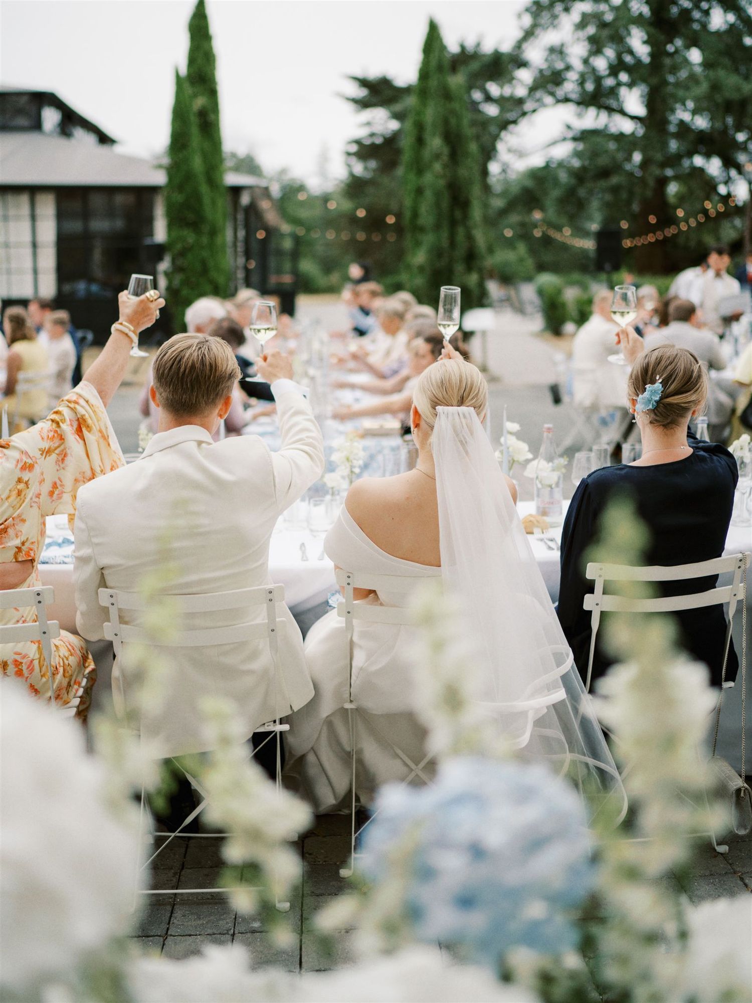 Wedding guests raising glasses in toast at elegant outdoor reception with floral arrangements.