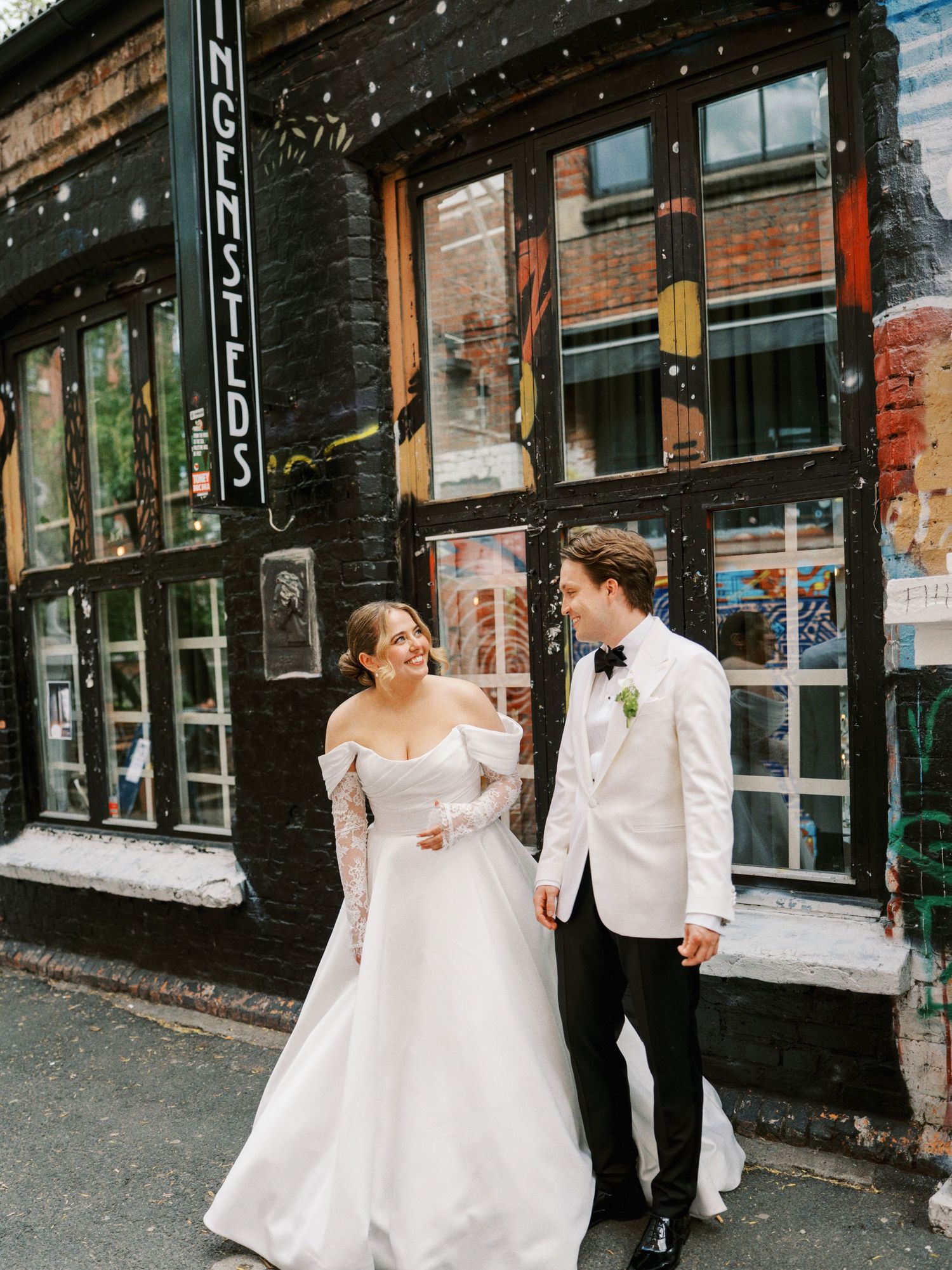 Bride and groom walking together outside a black brick building, smiling at each other on their wedding day.