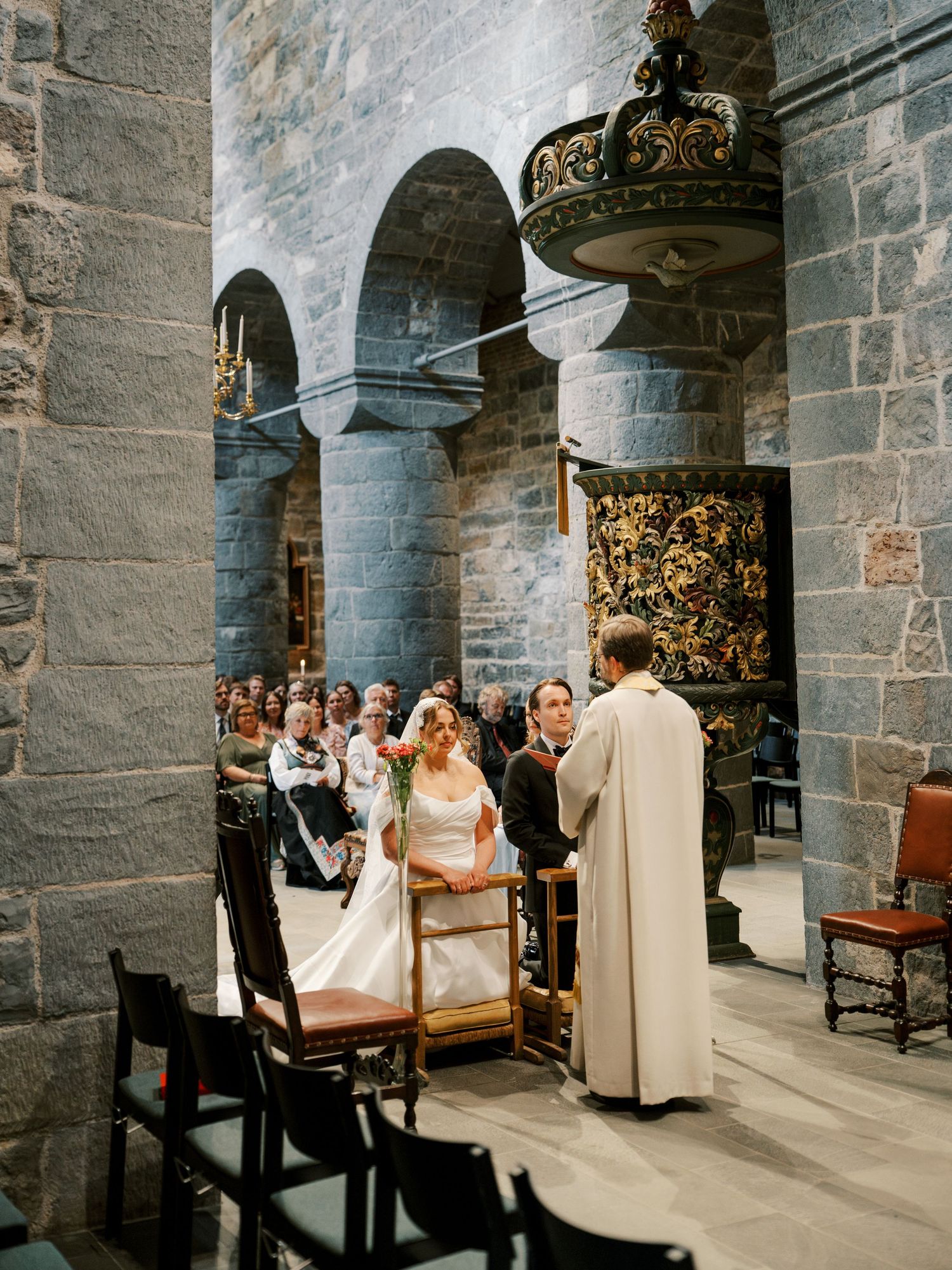 Bride and groom exchange vows at stone church altar with officiant, guests seated in historic cathedral.