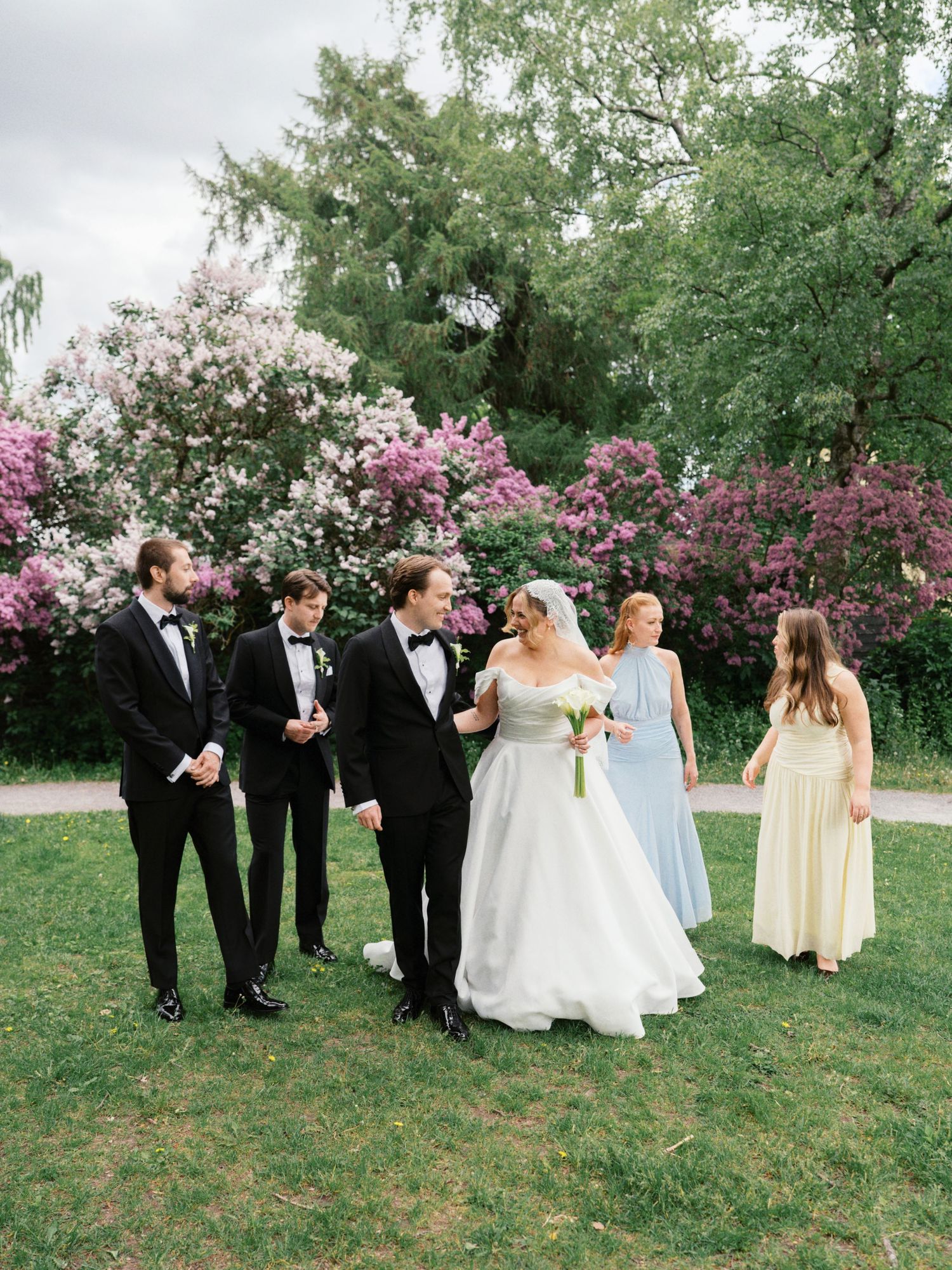 Wedding party walks on grass near blooming lilac bushes, bride in white gown with groom and guests in formal attire.