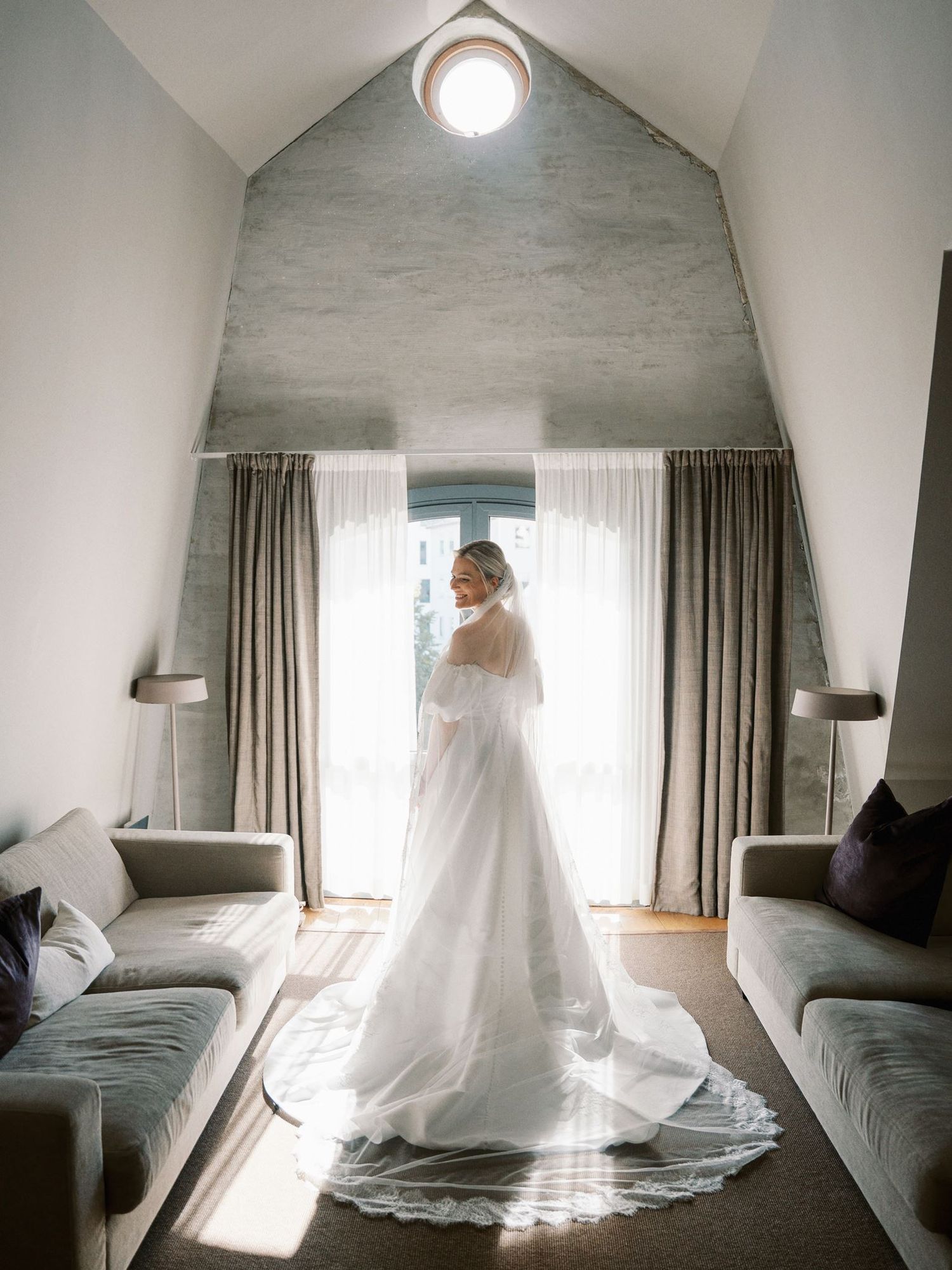 Bride in elegant white wedding gown with long train stands backlit by window in modern vaulted room.