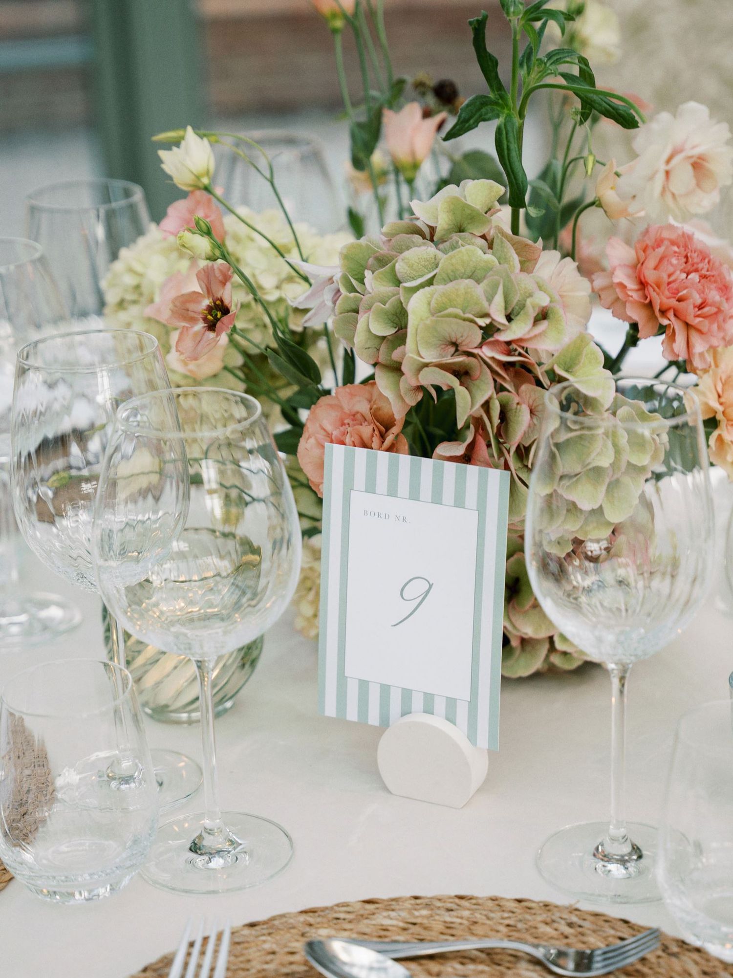 Elegant wedding table setting with pink and green floral centerpiece, crystal wine glasses, wicker chargers, and striped table number card.