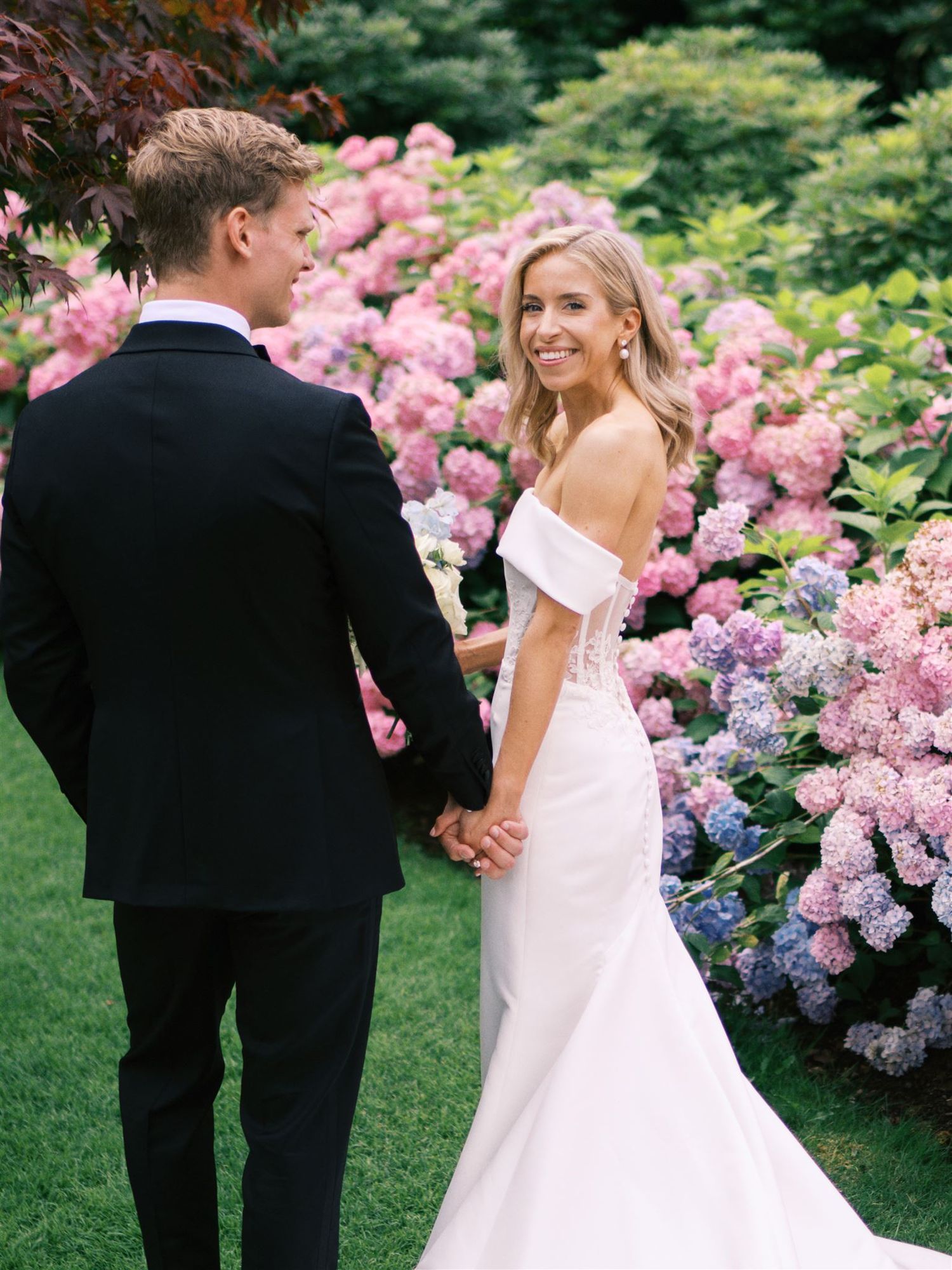 Bride in off-shoulder mermaid gown holds groom's hand before pink hydrangea garden.