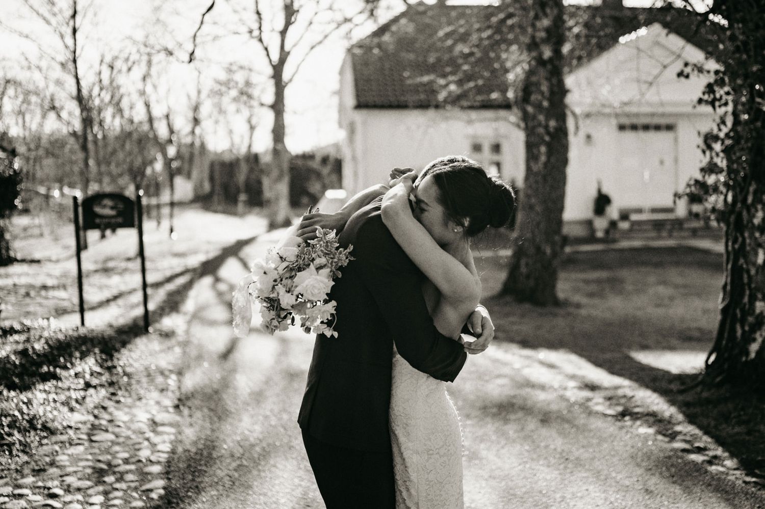 Black and white photo of bride and groom embracing on a tree-lined path outside a white country house.