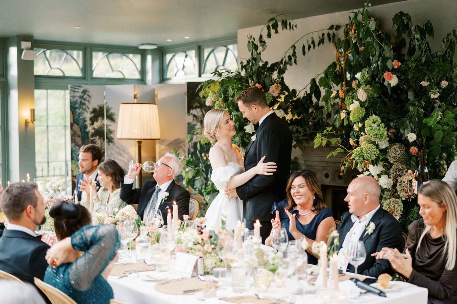 Bride and groom share a romantic moment at elegant wedding reception with lush floral backdrop and seated guests.