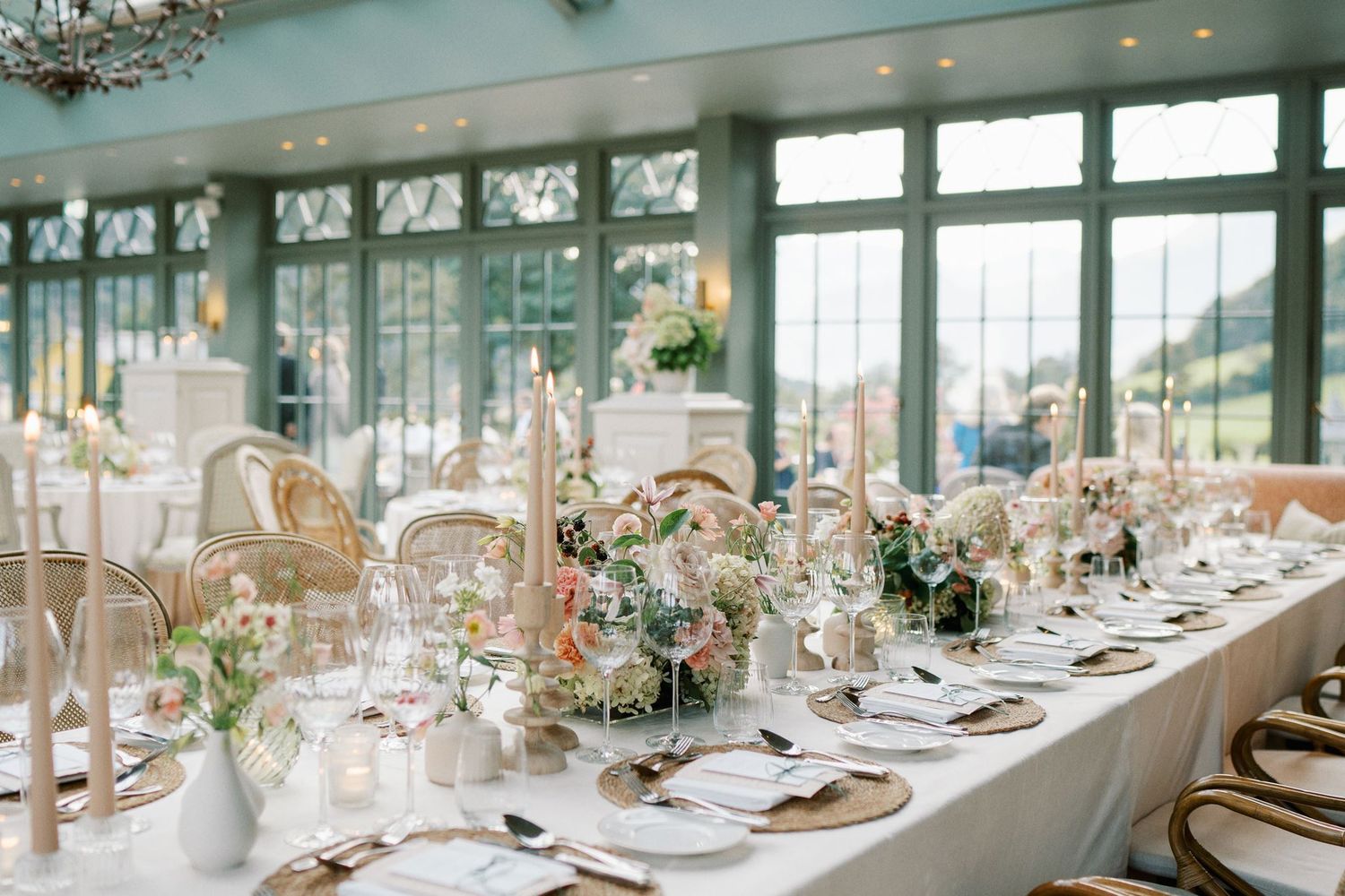 Elegant wedding reception with long white table, floral centerpieces, gold accents, and tall candles in a sunlit greenhouse venue.