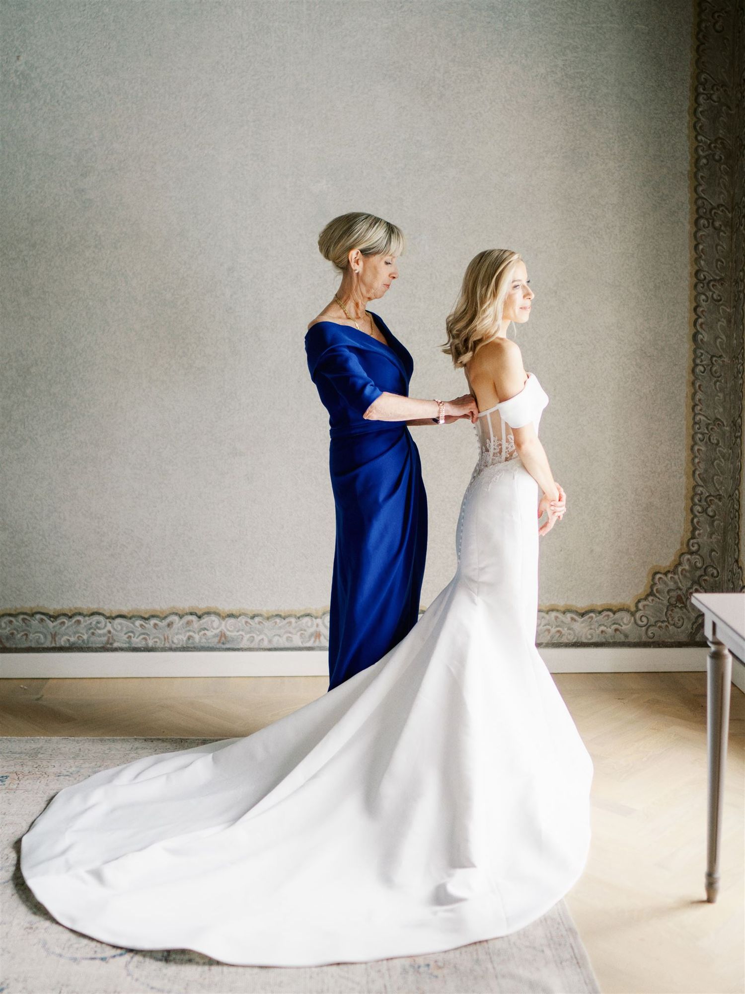 Mother in blue gown buttoning bride's white mermaid wedding dress in elegant room.