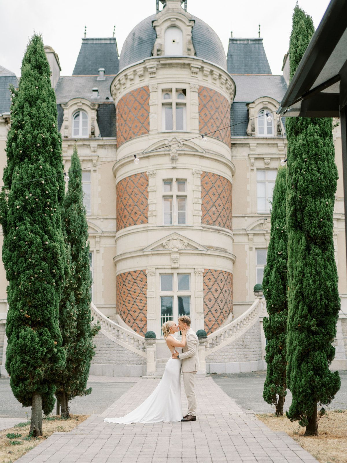 Bride and groom embracing in front of a grand French chateau with tall cypress trees.