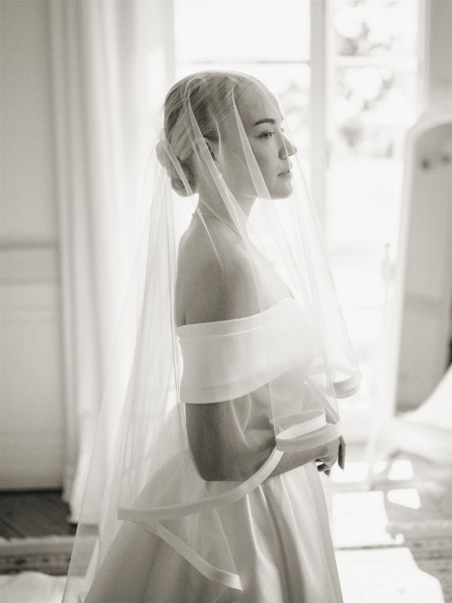 Elegant bride in off-shoulder gown and flowing veil posing by window in black and white.