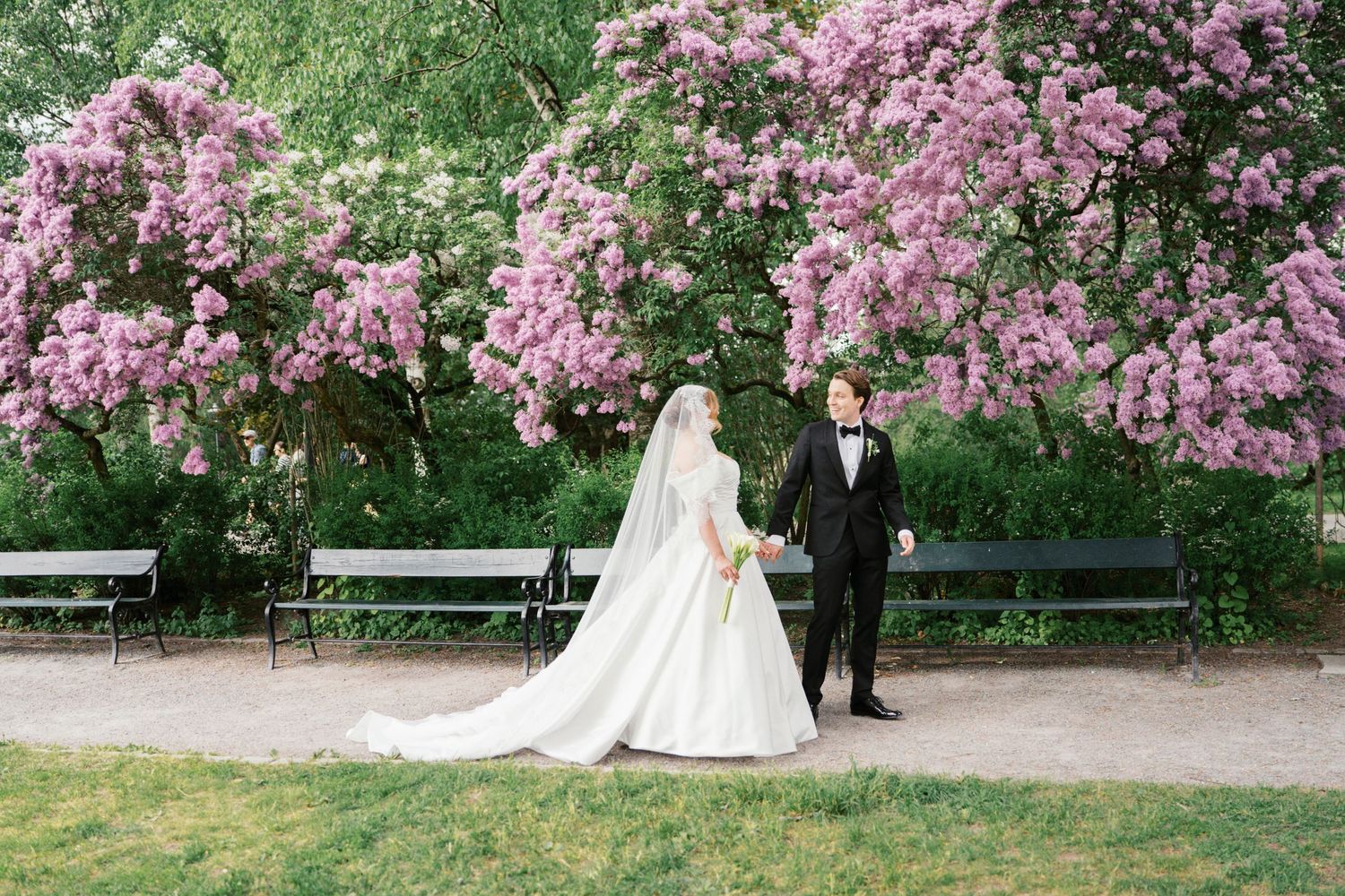A bride and groom hold hands near a blooming pink cherry blossom tree in a lush garden park setting.