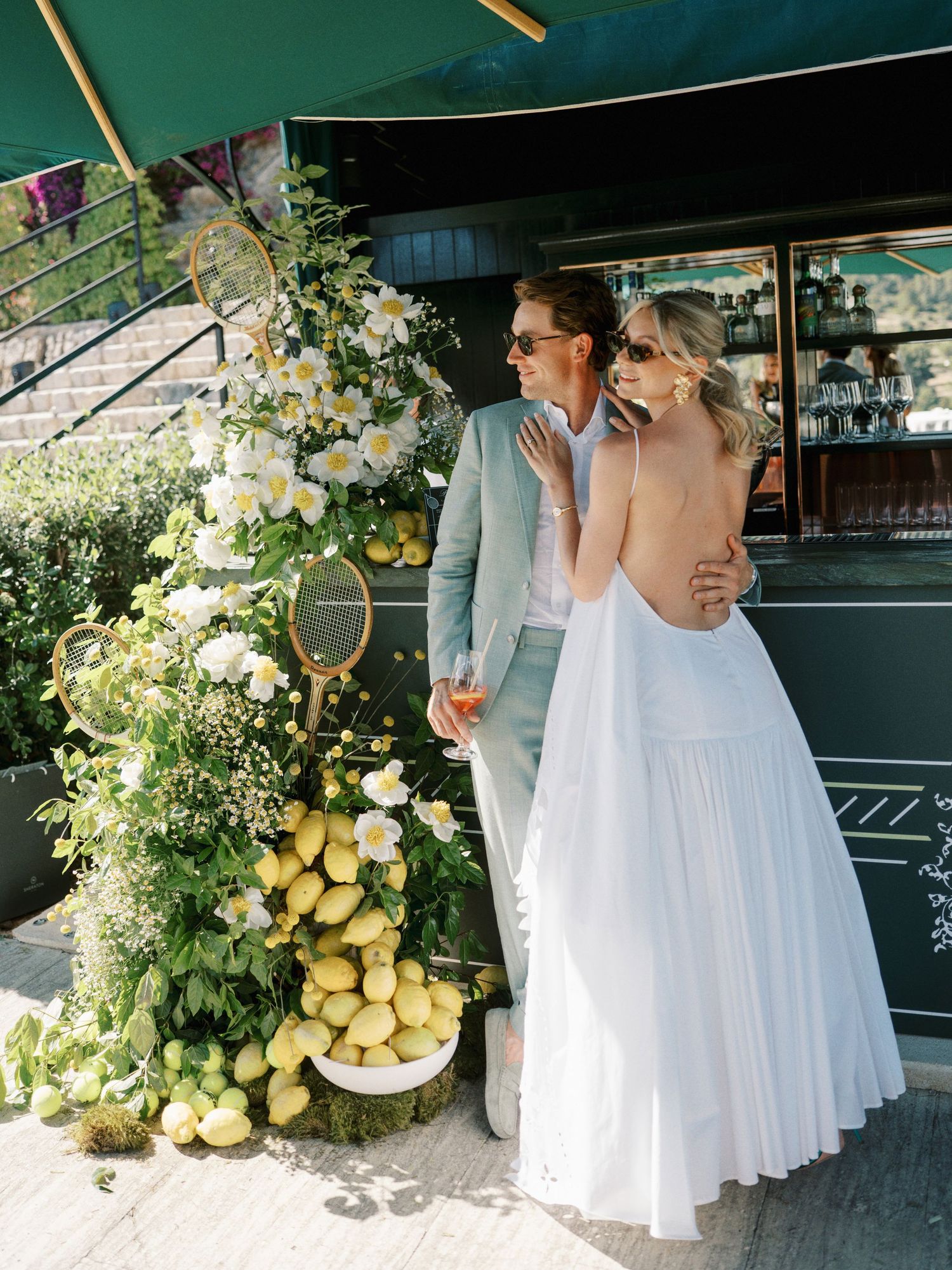 Bride in backless white gown and groom in light blue suit pose by lemon floral display at outdoor wedding venue.